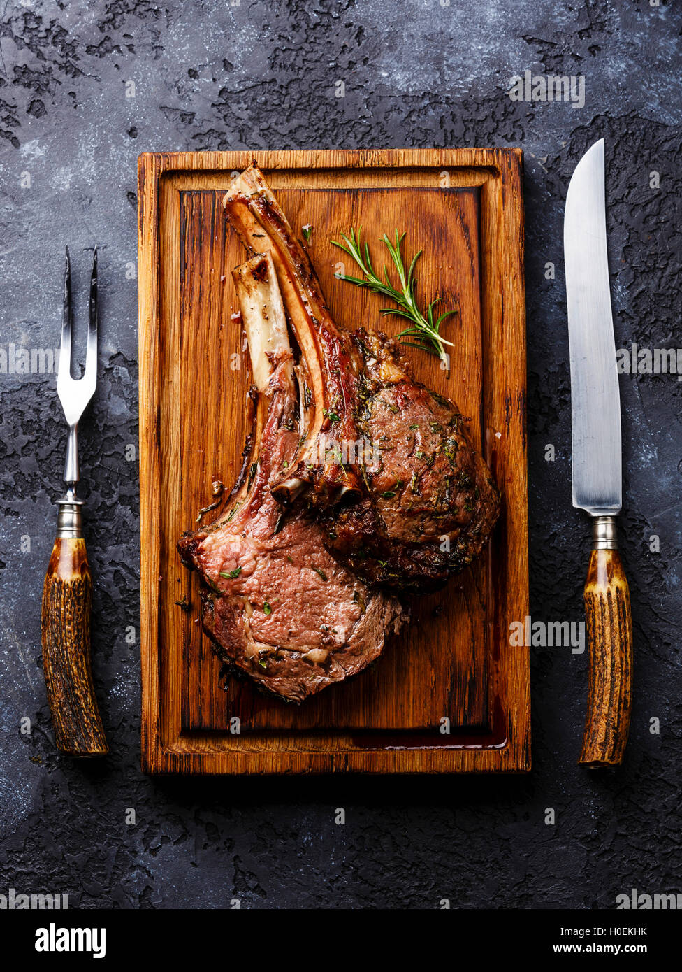 Arrosto di manzo nervature su osso sul tagliere di legno con coltello e forchetta carving impostato su sfondo scuro Foto Stock Arrosto di manzo nervature su osso sul tagliere di legno con coltello e forchetta carving impostato su sfondo scuro Foto Stock