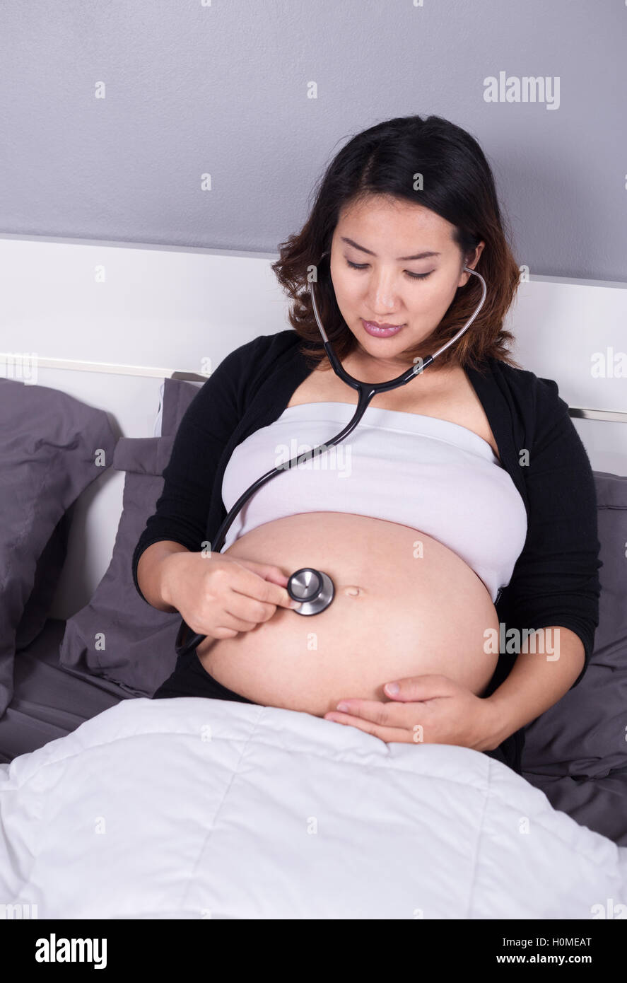 Donna incinta ascoltando il suo ventre con uno stetoscopio sul letto Foto Stock