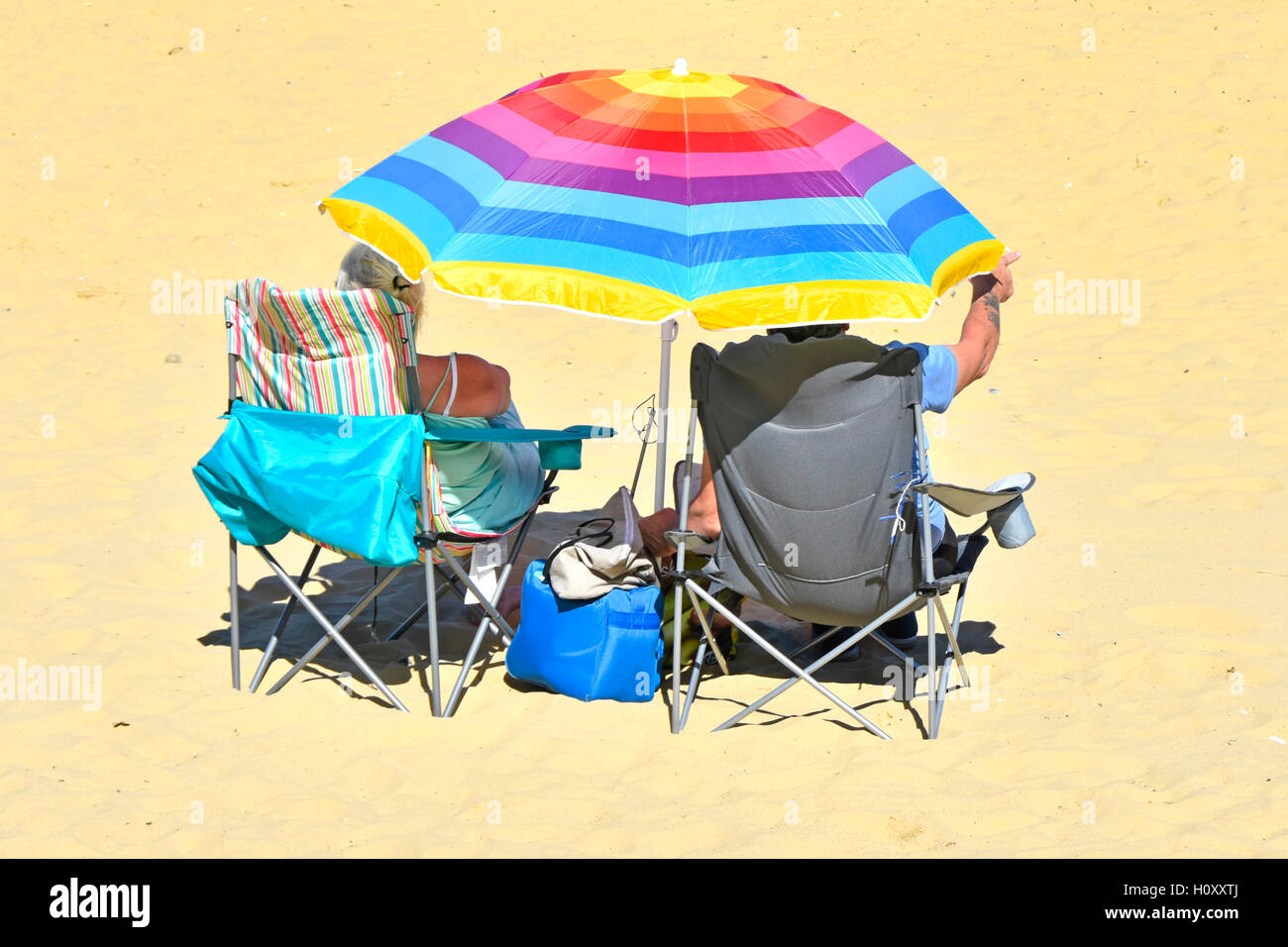 Coppia senior sotto ombrello colorato stile di vita rilassante ripiegare sedie spiaggia sabbiosa vacanza al mare britannico all'ombra dal caldo sole estivo UK Foto Stock
