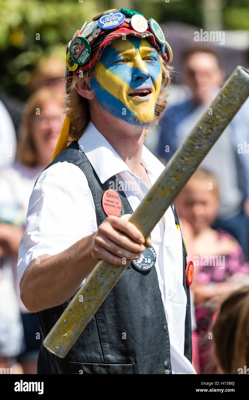 Chiusura del Royal Liberty Morris ballerino, giovane, con il volto dipinto di giallo e blu, indossa cappuccio rosso coperto di badge, scherma con palo metallico. Foto Stock