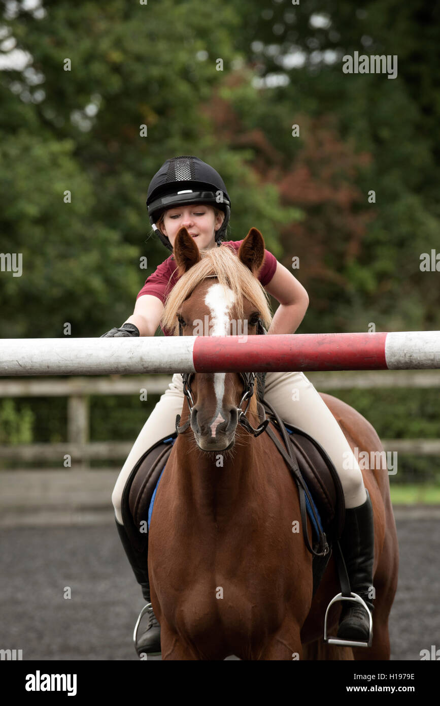 Giovane pilota che mostra un pony un palo alto su un salto - ragazza a cavallo di un pony di castagno che mostra l'animale l'altezza del salto Foto Stock