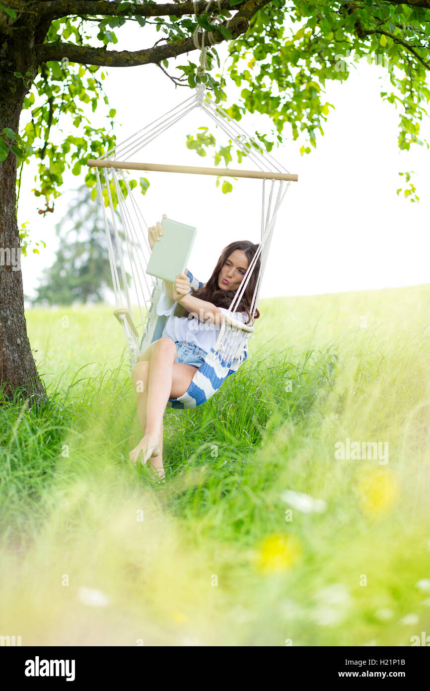 Donna prendendo selfie con la compressa in una sedia appeso sotto un albero Foto Stock