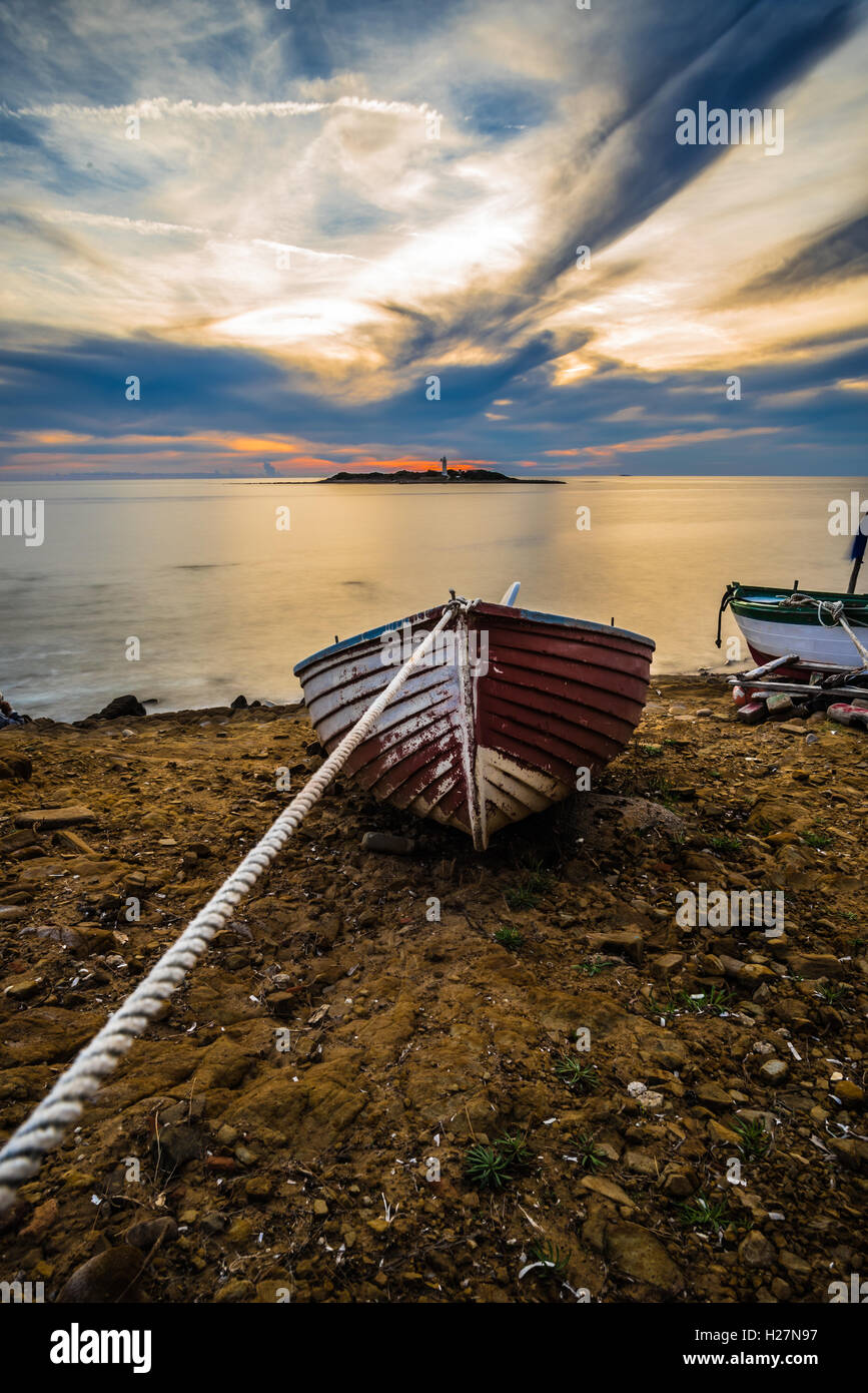 Una fune tiene una vecchia imbarcazione in legno attratti verso il mare piatto, il tramonto e le nuvole in movimento. Foto Stock