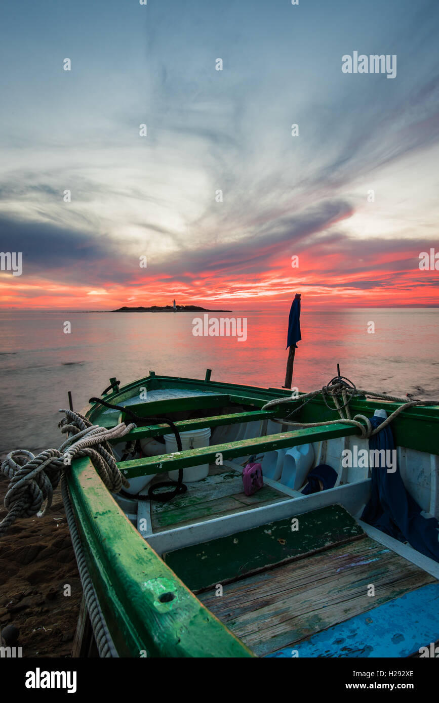 Vecchia barca in legno, funi, piatto mare, un'isola con il faro sotto un tramonto cotto Foto Stock
