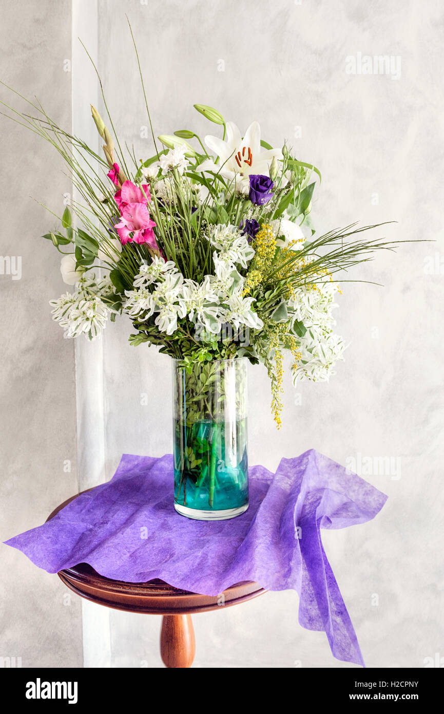 Colorate composizioni floreali con gigli, rose e vegetazione in un vaso di vetro su un tavolo, accentuate da tessuto viola contro una parete luminosa. Foto Stock