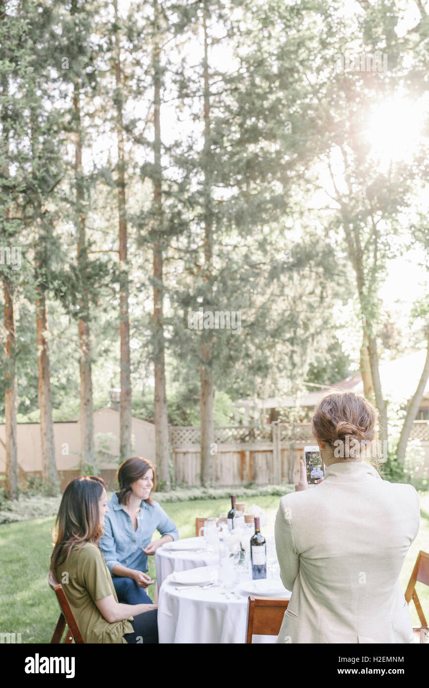 Donna che utilizza il suo telefono cellulare, scattare una fotografia di un gruppo di donne sedute a un tavolo in un giardino. Foto Stock