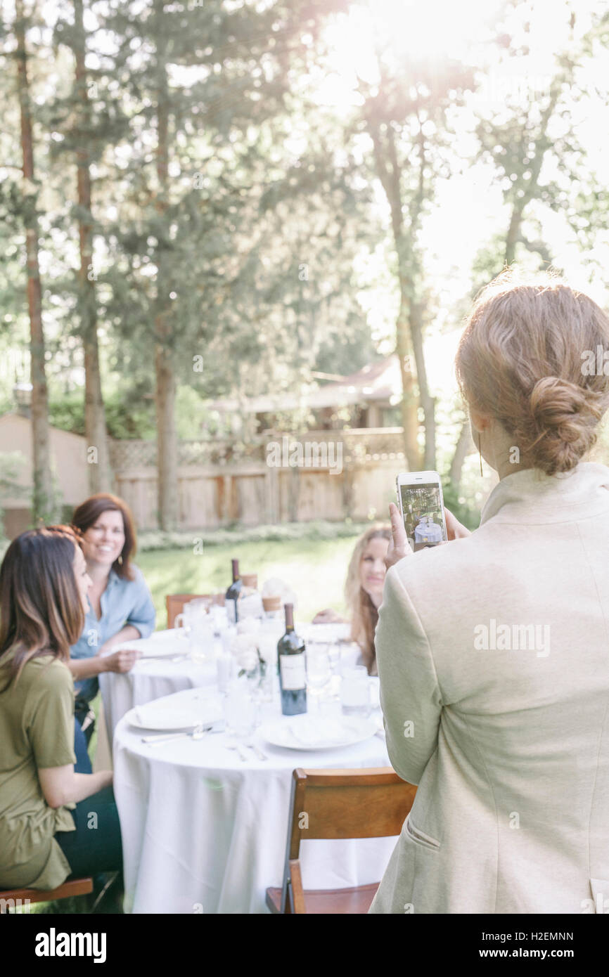 Donna che utilizza il suo telefono cellulare, scattare una fotografia di un gruppo di donne sedute a un tavolo in un giardino. Foto Stock