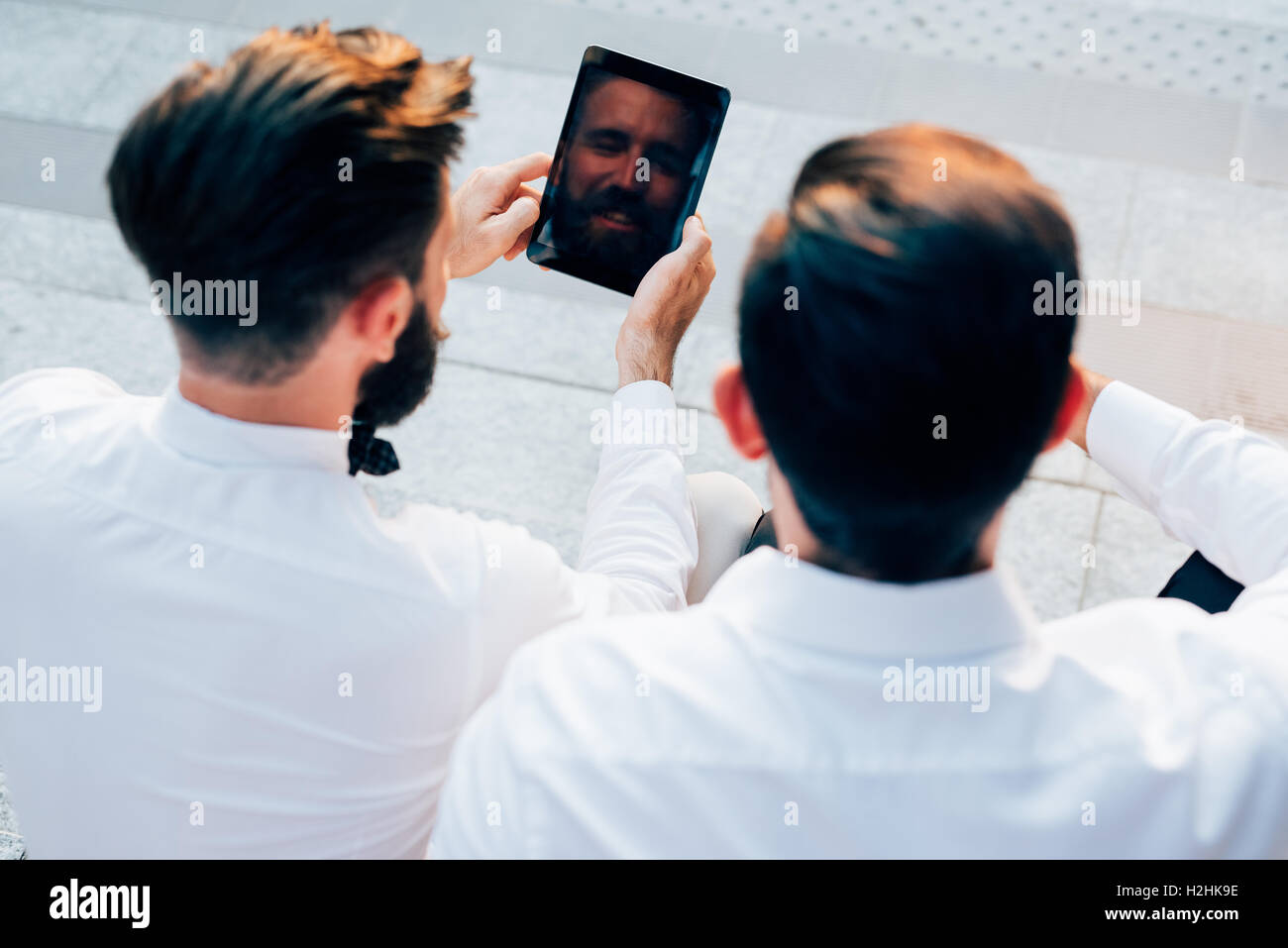 Vista da dietro di due giovani barbuto bionda e capelli neri imprenditore moderno, seduti su una scalinata, utilizzando tablet - technolo Foto Stock