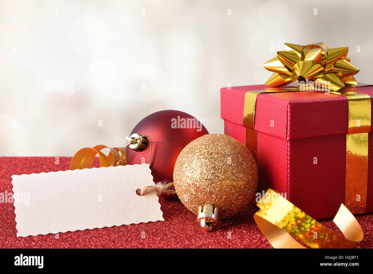 Decorazione di natale con tag, confezione regalo rossa e nastro d'oro e due sfere sul rosso brillante tabella strutturata e sfondo bianco. Parte anteriore Foto Stock