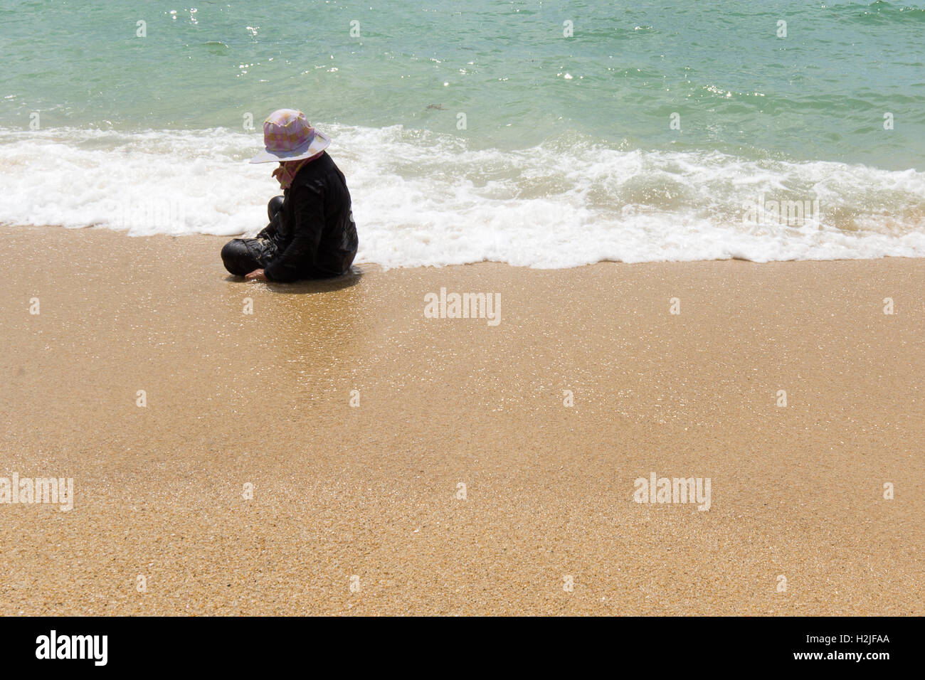 Thai donna seduta sulla spiaggia in un abito nero e un grande cappello, picking cozze in terra-wash. Spazio per testi Foto Stock