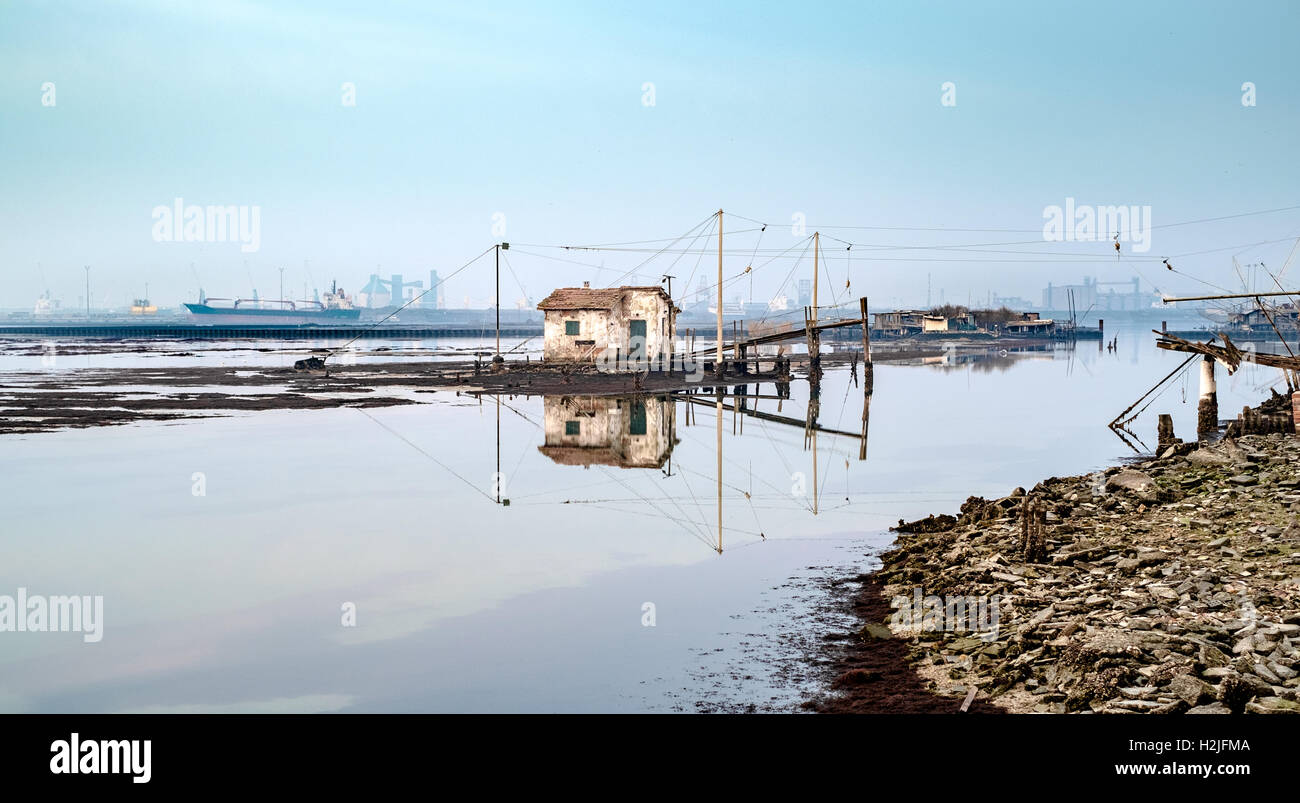 Vecchia casa di pesca che riflette nella laguna di Marina di Ravenna, porto industriale sullo sfondo. Foto Stock