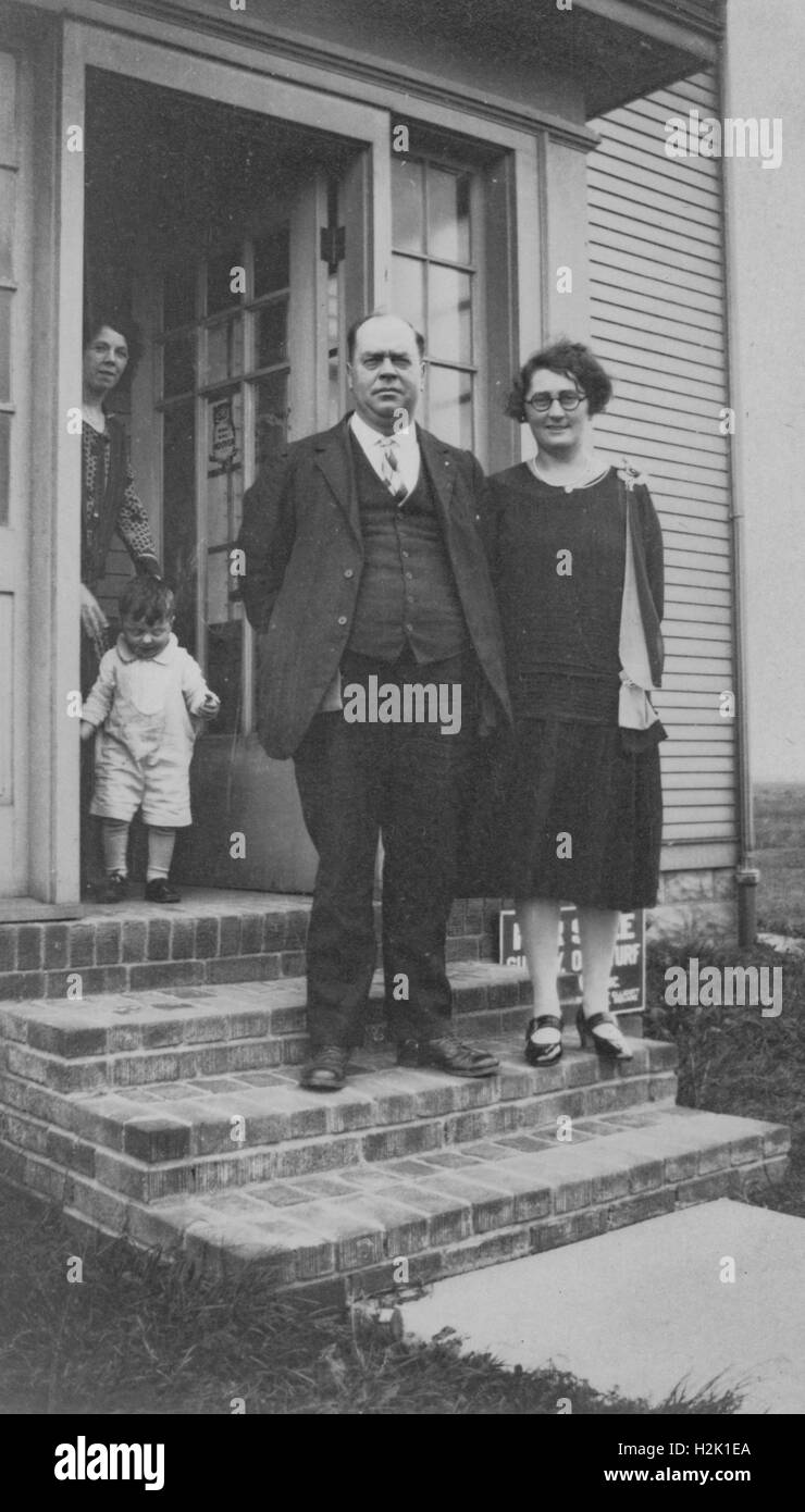Unknown City, Stati Uniti - Gennaio 01, 1950: una coppia matura stand sulla parte anteriore fasi della loro casa con una giovane donna e bambino visibile all'interno dello sportello anteriore, 1950 Foto Stock