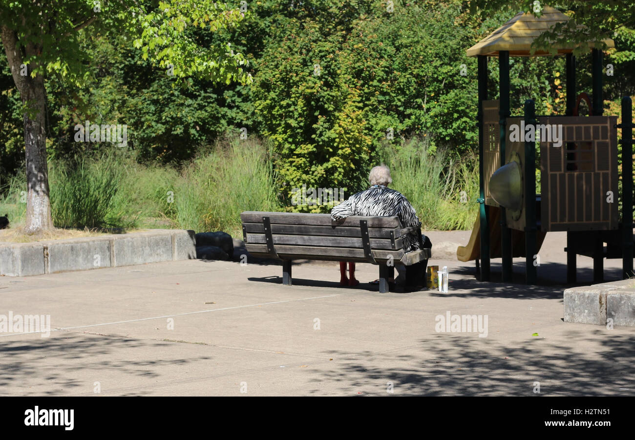 Donna anziana seduta su una panchina nel parco vicino a giochi per bambini struttura, acqua da bere e mangiare crackers di sunny estate Foto Stock