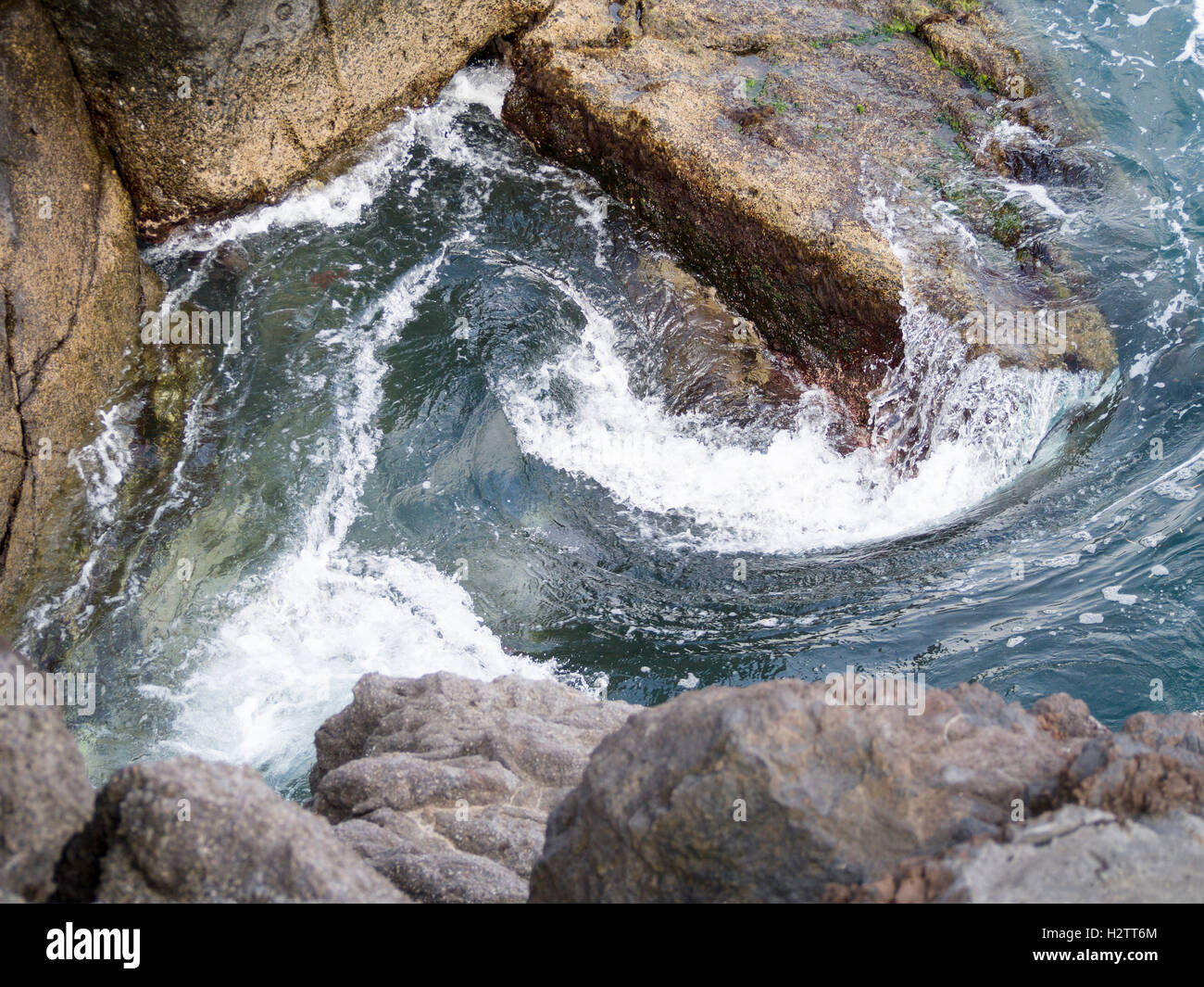 Surf vorticoso intorno alle rocce. L'acqua dalle onde colpire la costa rocciosa e curl in una vasca idromassaggio di surf. Foto Stock