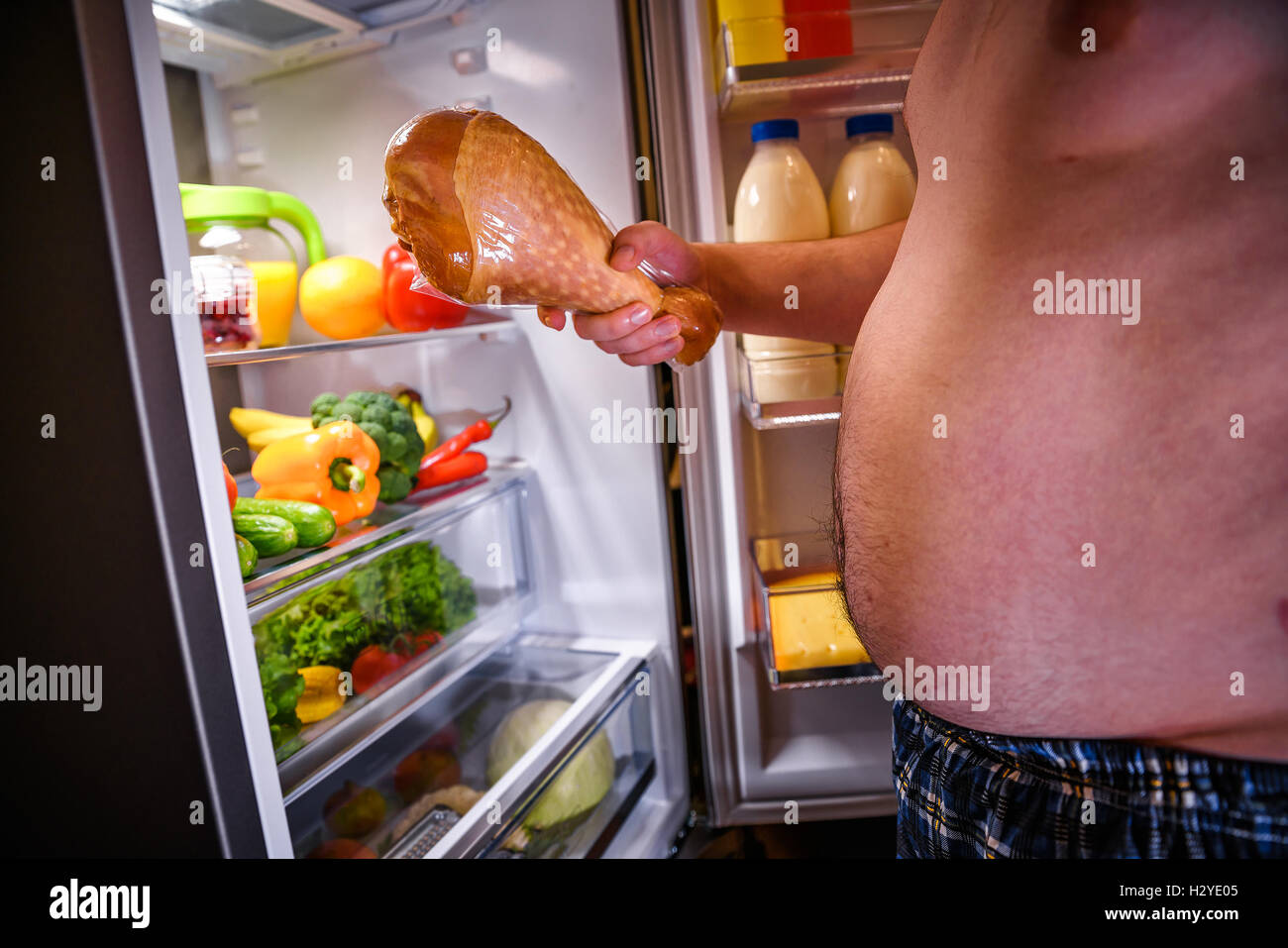 Fame fat man tenendo un grosso tacchino nelle sue mani e piedi accanto al frigorifero aperto. Alimenti dannosi per la salute. Foto Stock