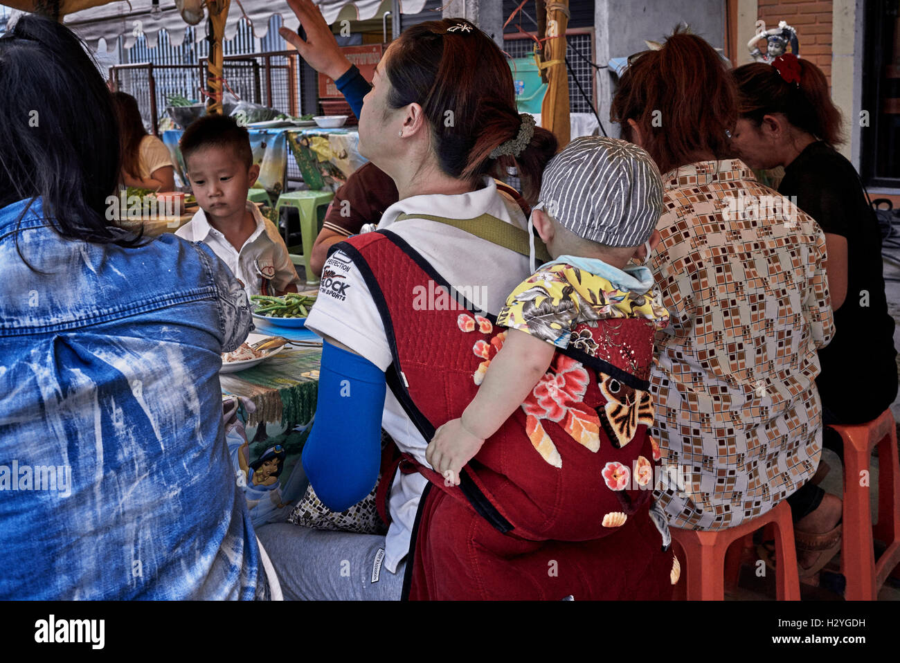 Madre con bambino portato in un imbragatura posteriore e mangiare in un ristorante all'aperto. Thailandia S. E. Asia Foto Stock