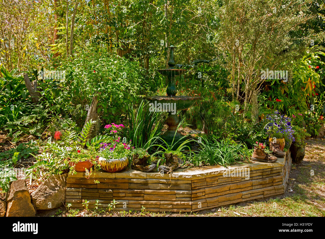 Giardino ornato di fontana circondata da fogliame color smeraldo, alberi & bassa parete di roccia con una decorazione di contenitori di fiori colorati Foto Stock