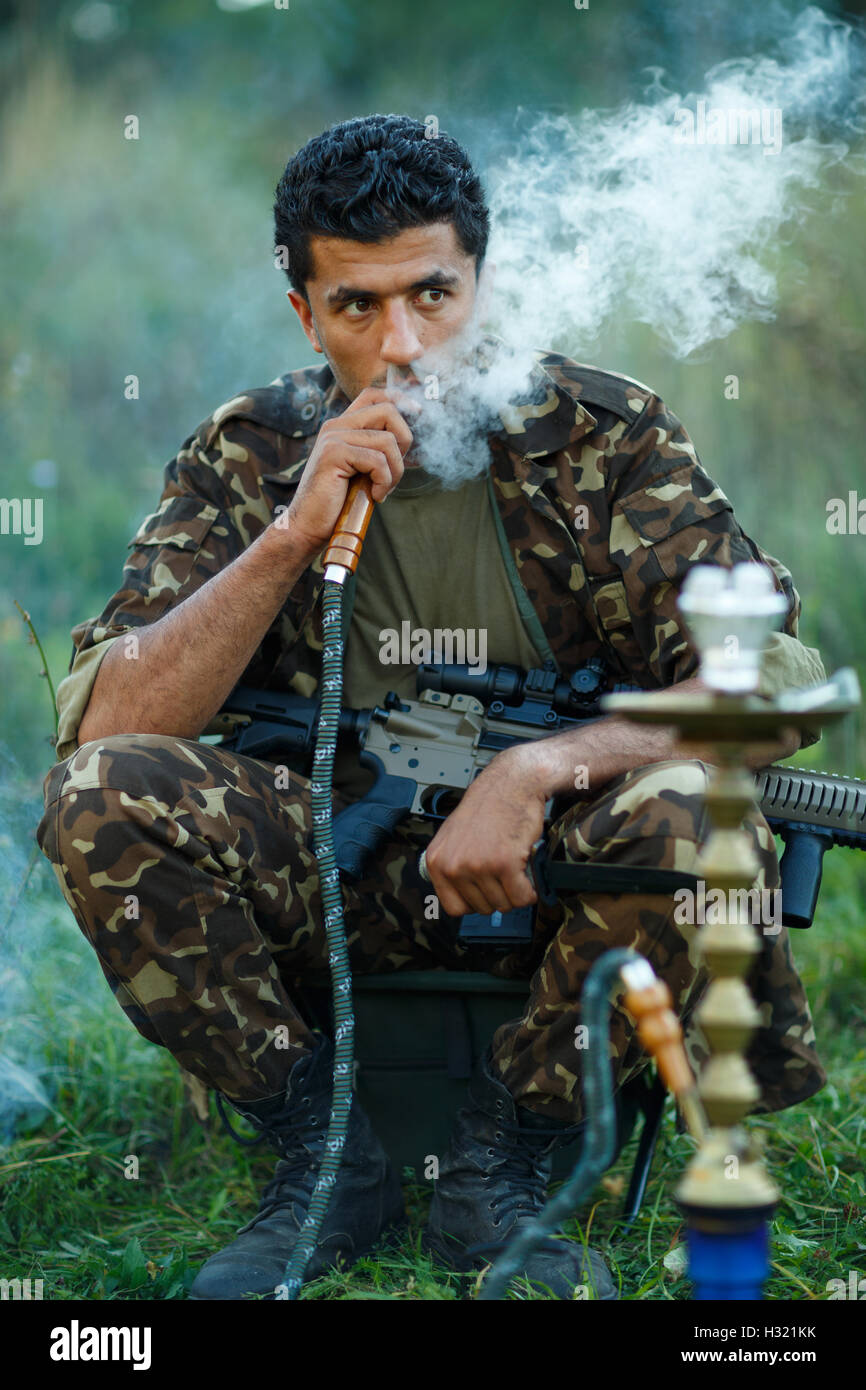 Uomo di nazionalità arabo in camuffamento con un fucile da caccia fuma un narghilè outdoor Foto Stock