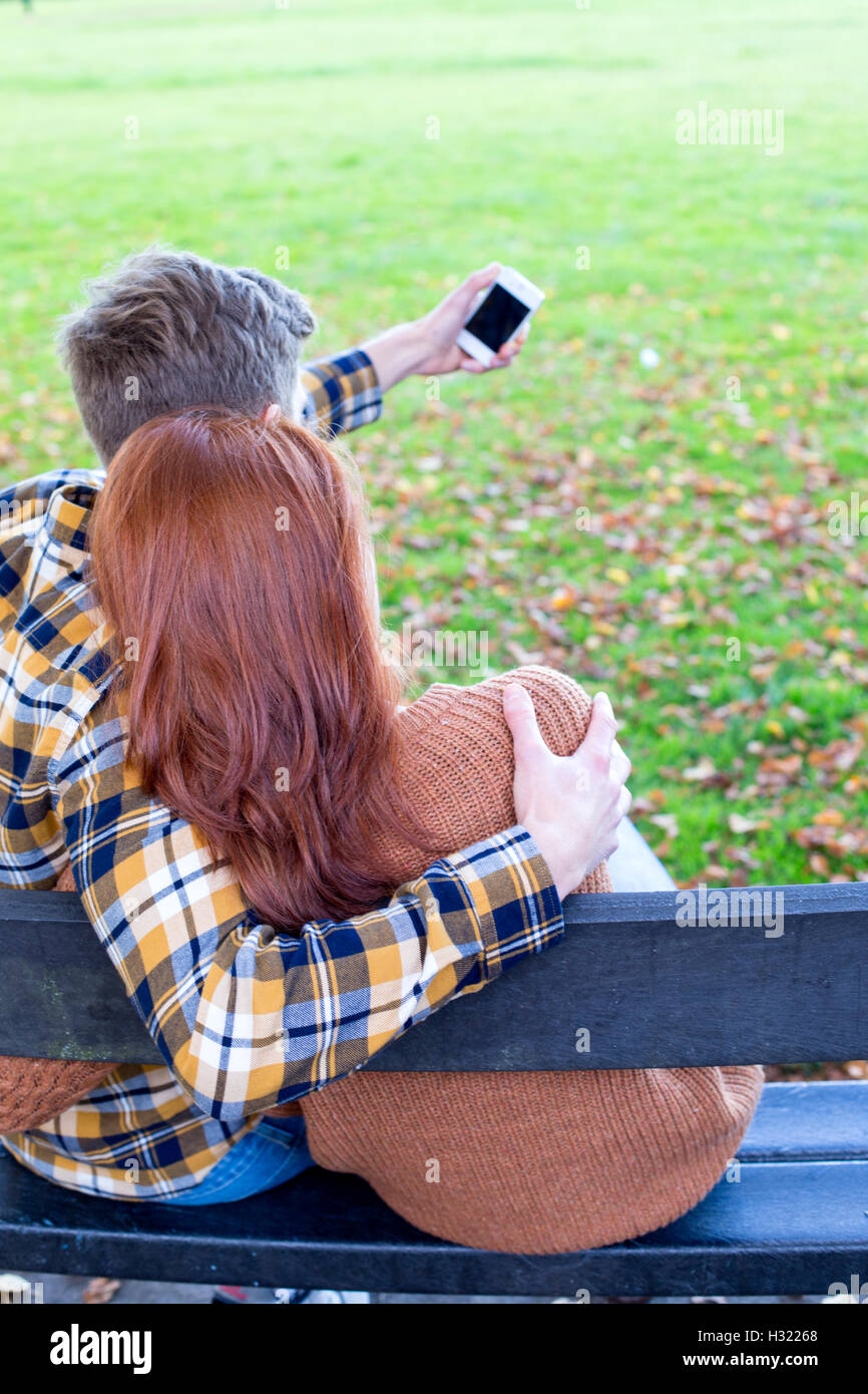 Vista posteriore di una giovane coppia di prendere una selfie su uno smartphone. Essi sono coccole su un banco in un parco. Foto Stock