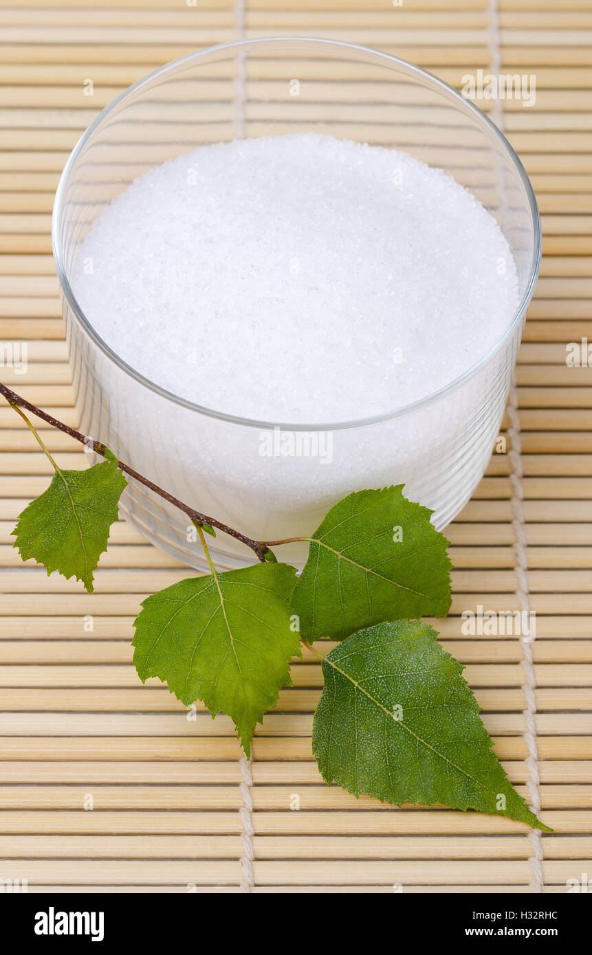 Xilitolo birch lo zucchero in un recipiente di vetro con foglie di betulla su un tappetino di bambù. Lo zucchero cristallizzato bianco alcol, un dolcificante. Foto Stock