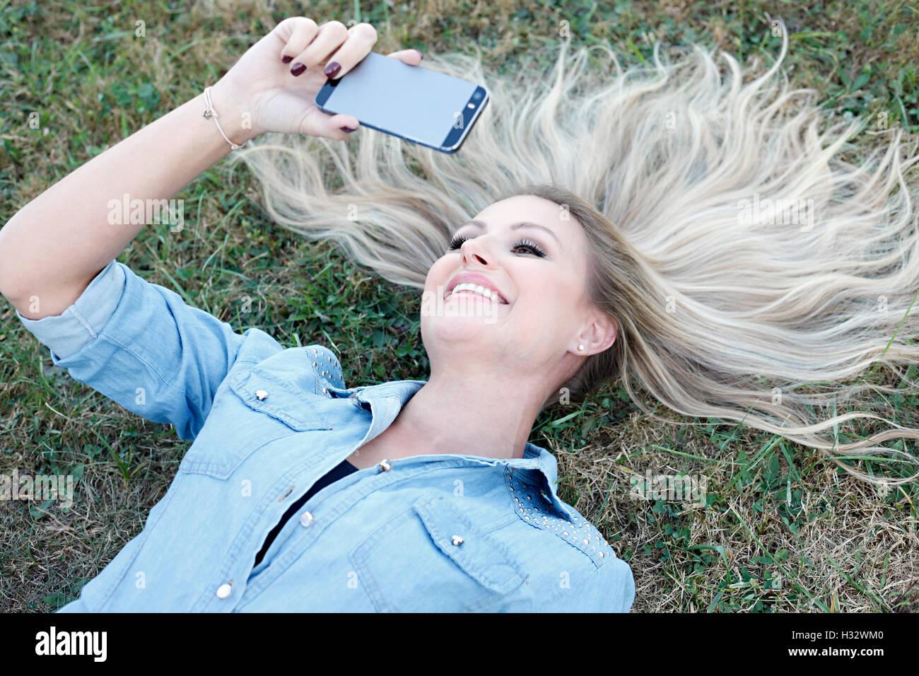 Giovane e bella bionda sdraiati sull'erba e tenendo selfie Foto Stock
