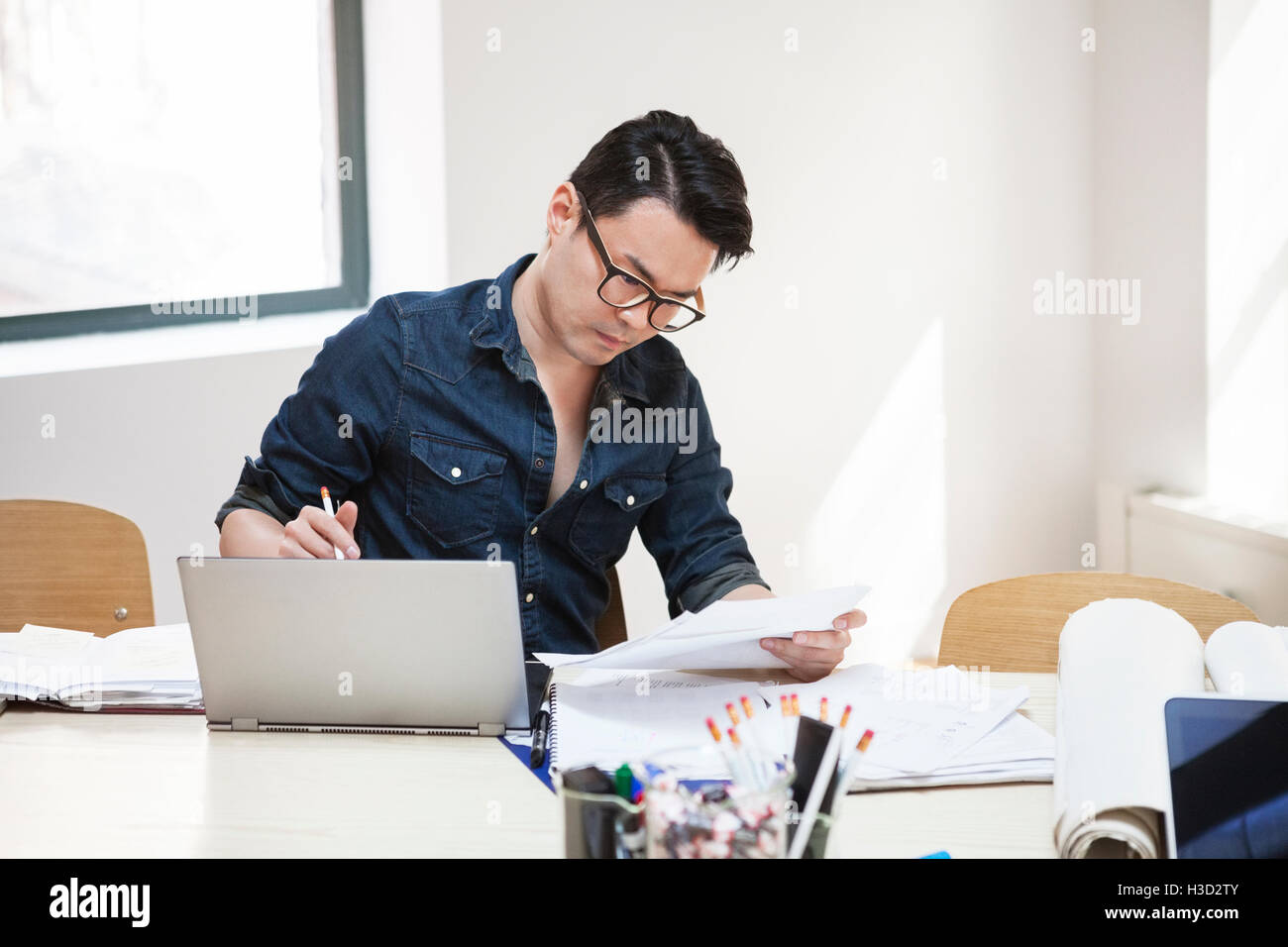 uomo d'affari che legge documenti mentre utilizza un computer portatile alla scrivania nell'ufficio creativo Foto Stock