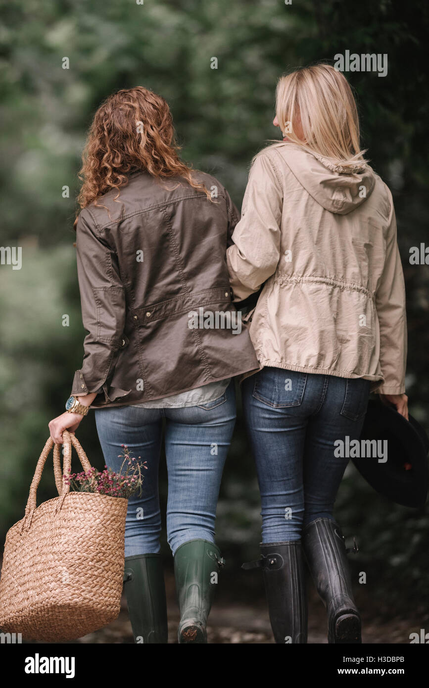 Due donne a braccetto a piedi in cappotti e stivali lungo un percorso di paese con un cestello. Foto Stock