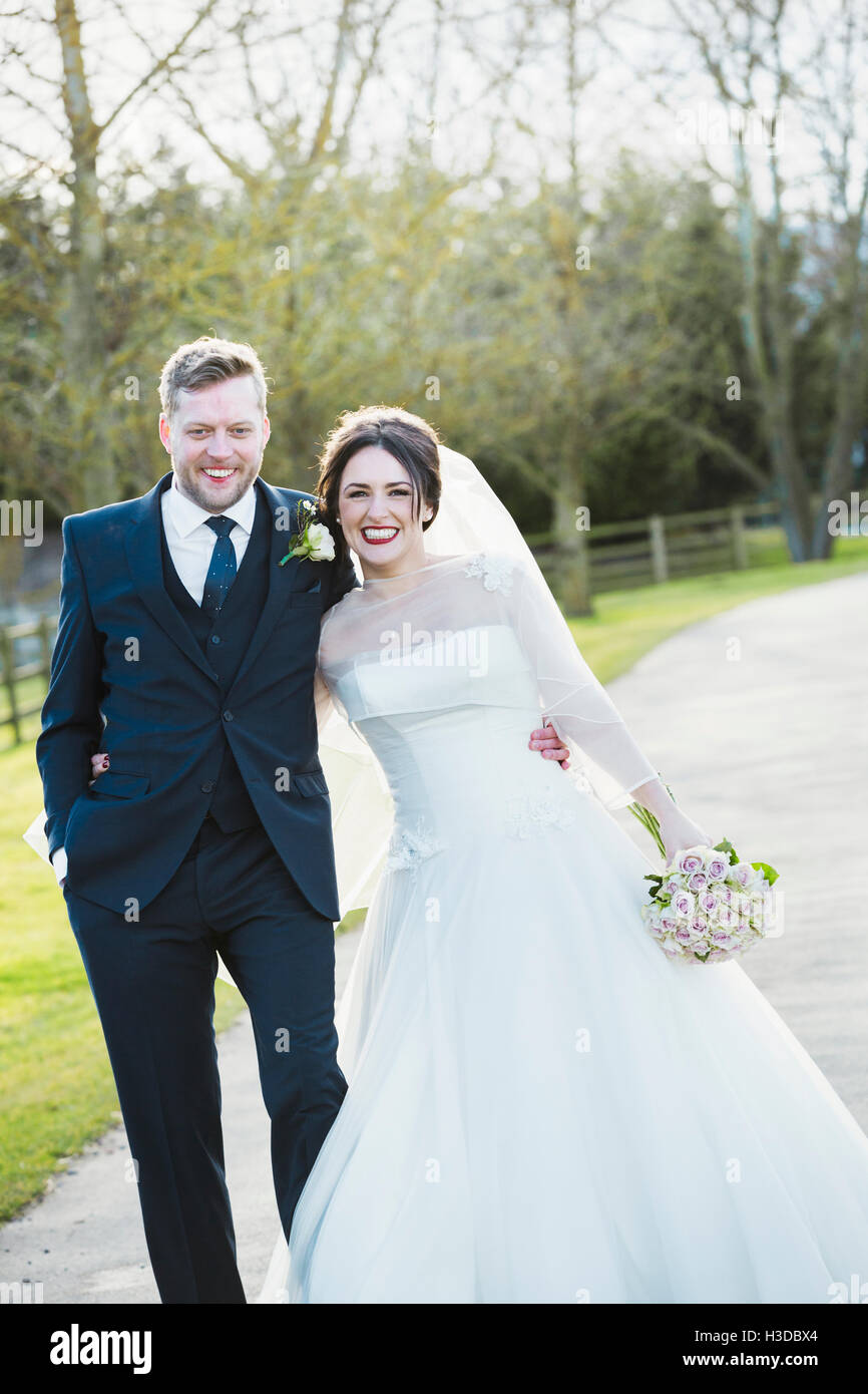 La sposa e lo sposo il giorno delle nozze camminando sotto braccio giù per un sentiero sotto il sole a ridere. Foto Stock