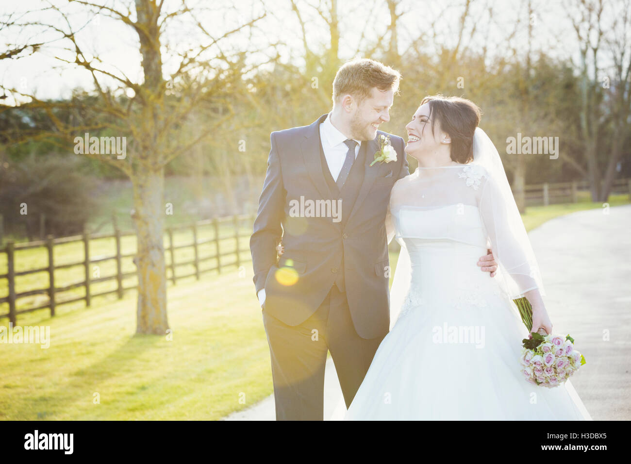 La sposa e lo sposo il giorno delle nozze camminando sotto braccio giù per un sentiero sotto il sole a ridere. Foto Stock