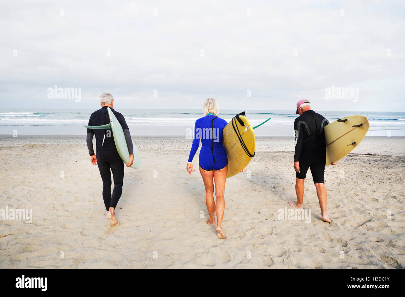 Donna Senior e due alti uomini su una spiaggia, indossa una muta e il trasporto di tavole da surf. Foto Stock