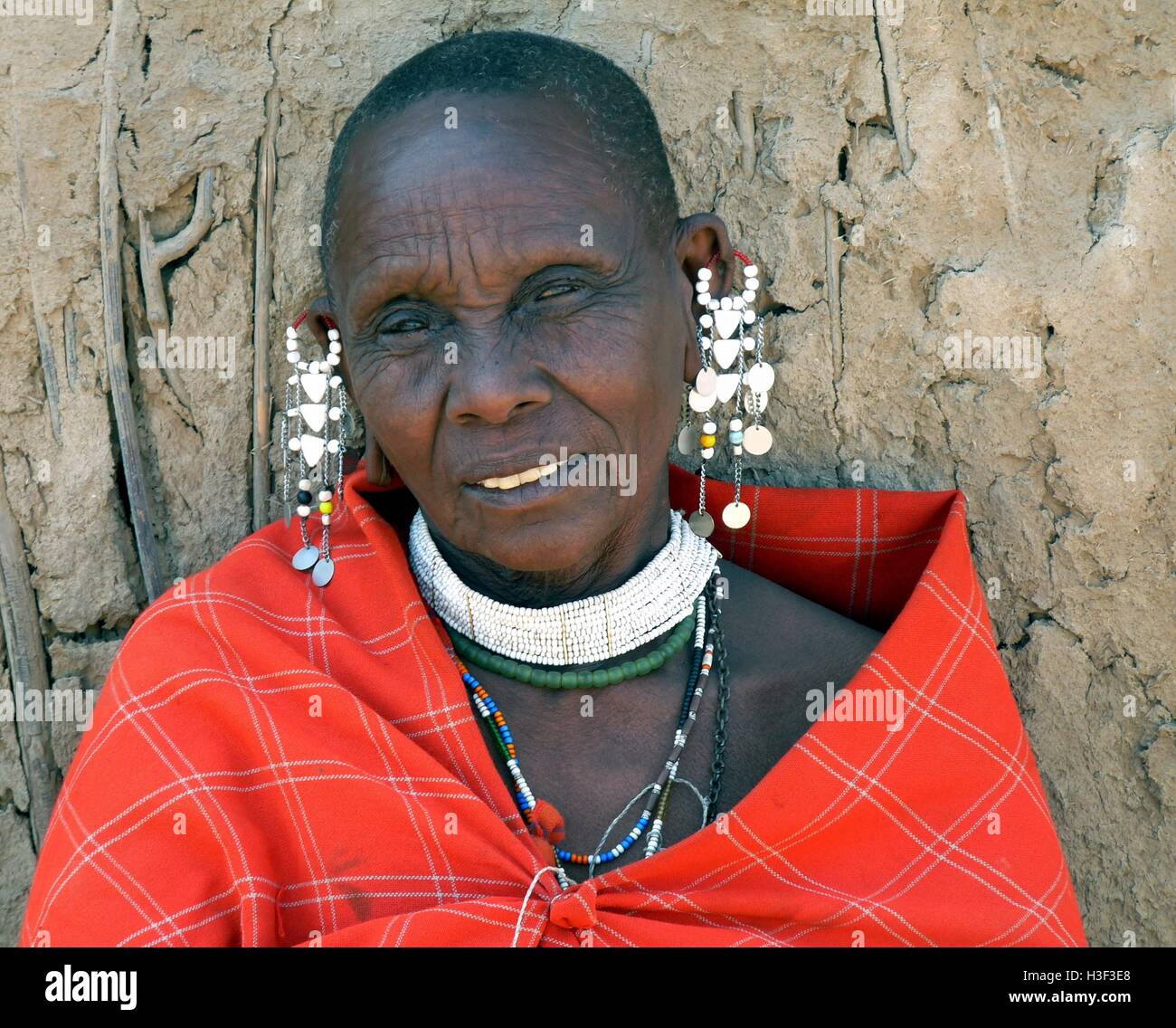 Close up di anziani Masai donna che indossa tradizionale rosso coperta e intricato fatti a mano gioielli di perline di fronte a capanna di fango Foto Stock