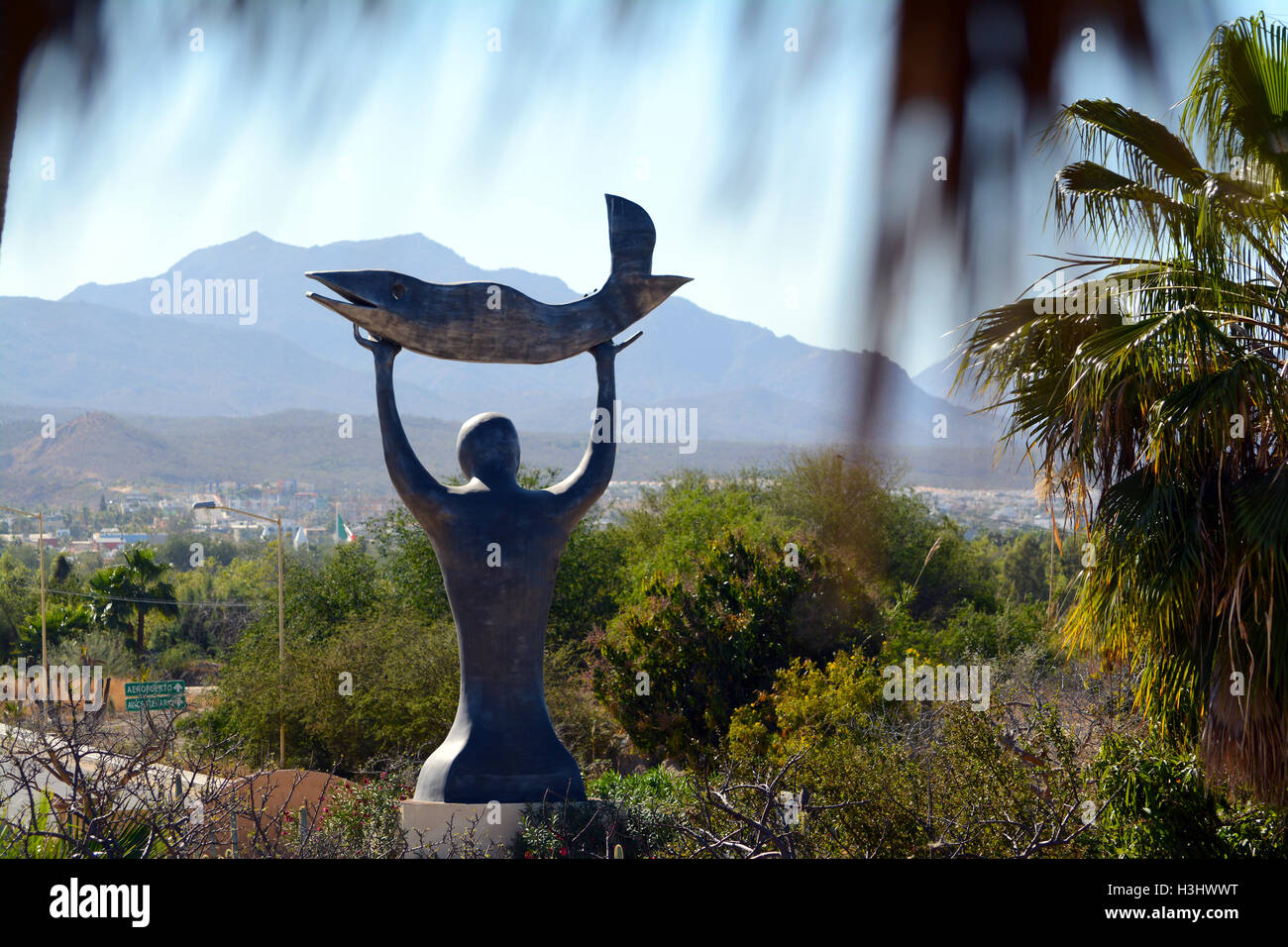 Statua di un uomo con un pesce verso il cielo a Wirikuta Giardino Botanico, Baja, Cabo, Messico con le montagne e il cielo blu Foto Stock