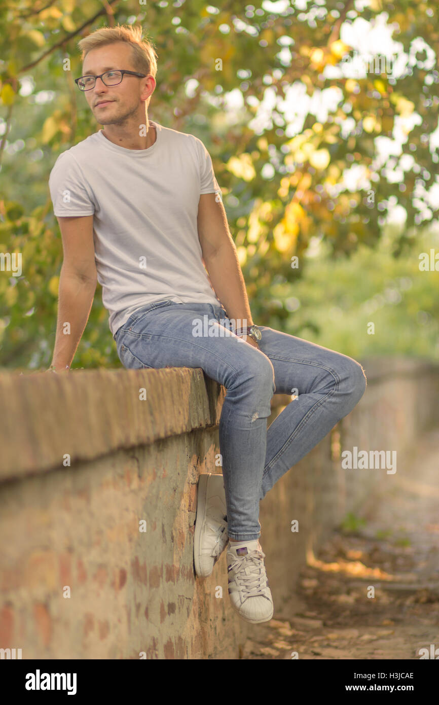 Giornata di sole un giovane uomo all'aperto estate seduta abiti casual t-shirt bianco jeans Foto Stock