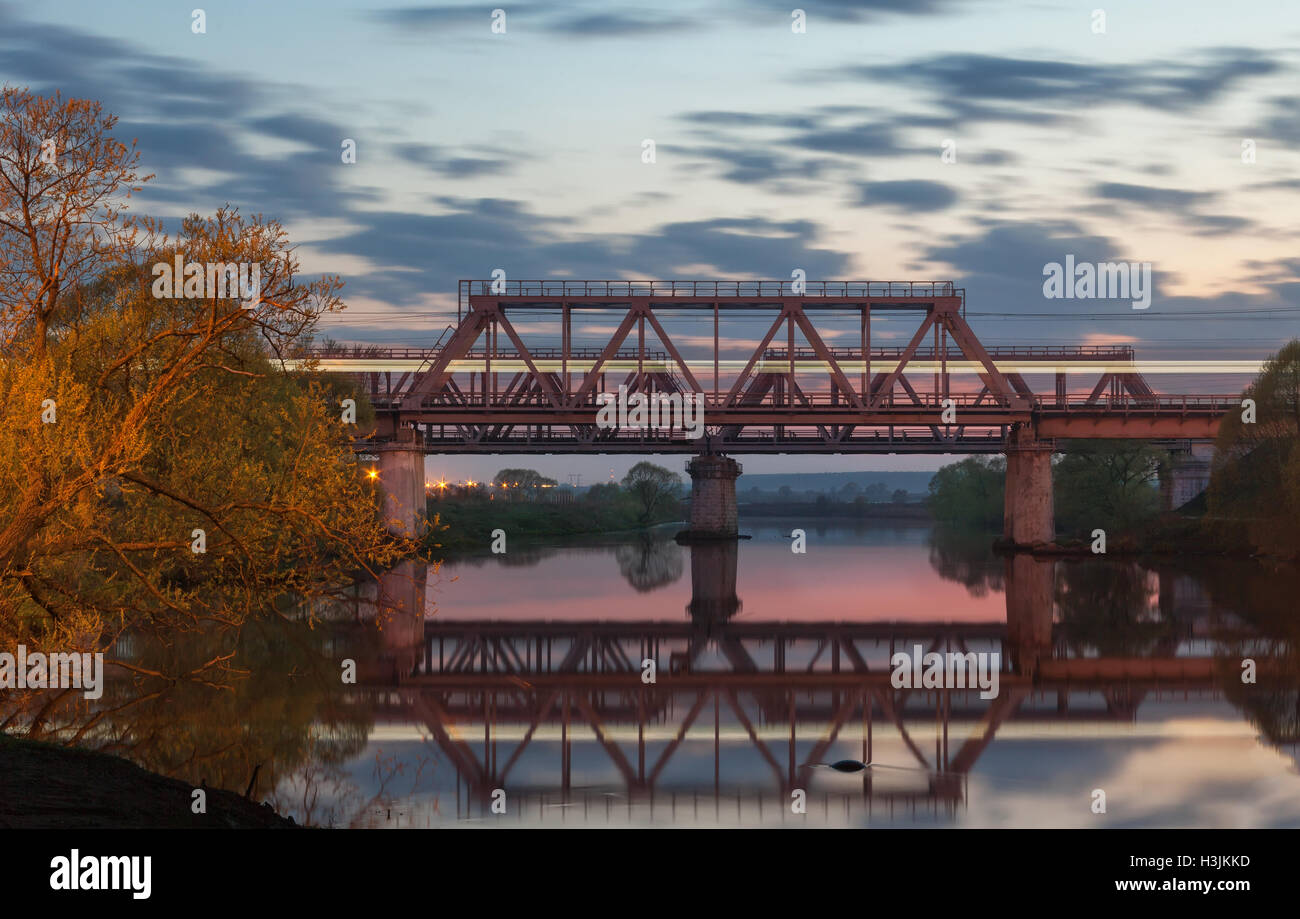 Ponte Ferroviario sul fiume al tramonto con luce sfocata su un treno passare Foto Stock
