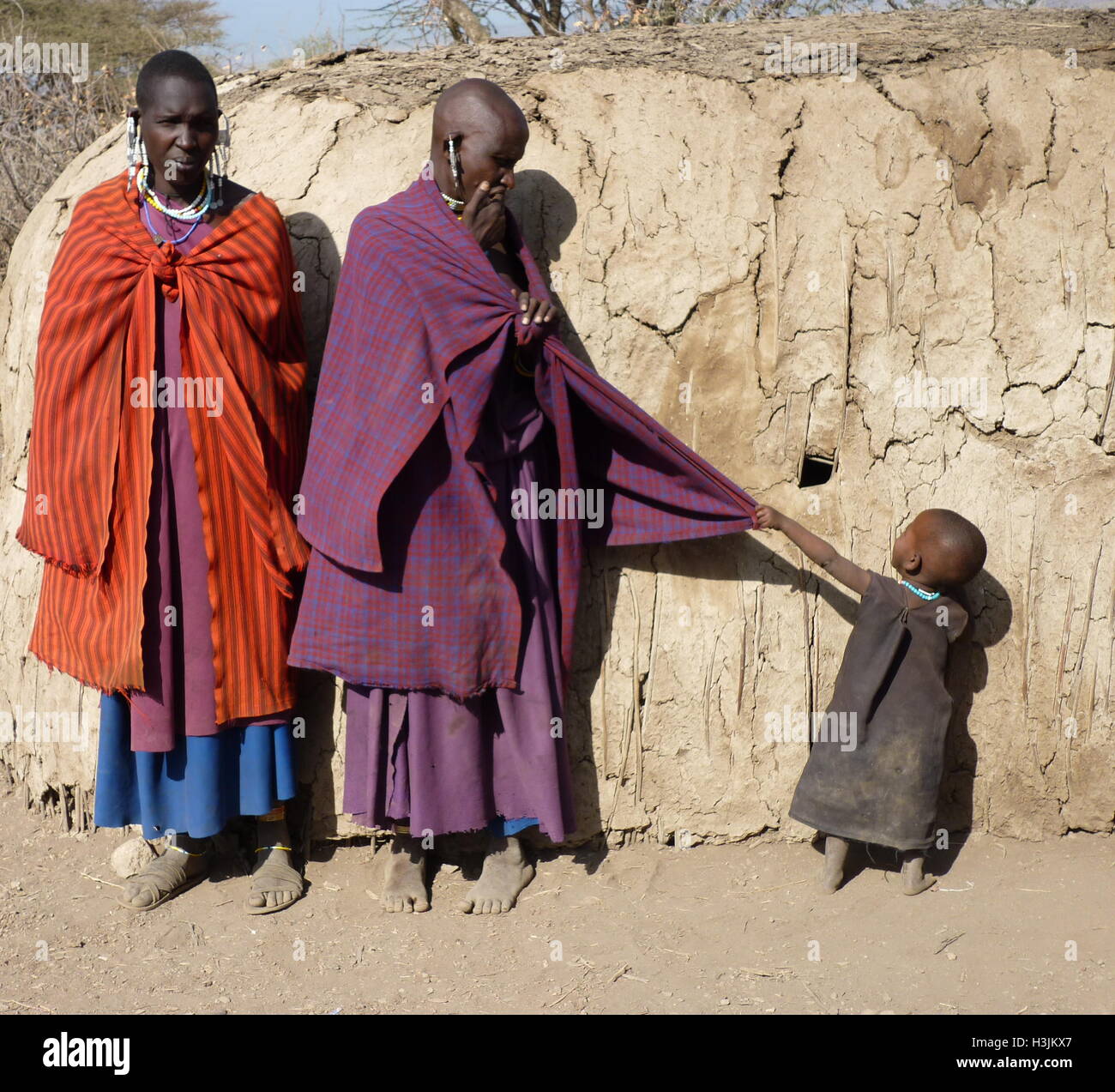 Masai famiglia in abiti tradizionali con un bambino piccolo tirando a madre di vestiti per ottenere attenzione,in piedi di fronte a capanna di fango. Foto Stock