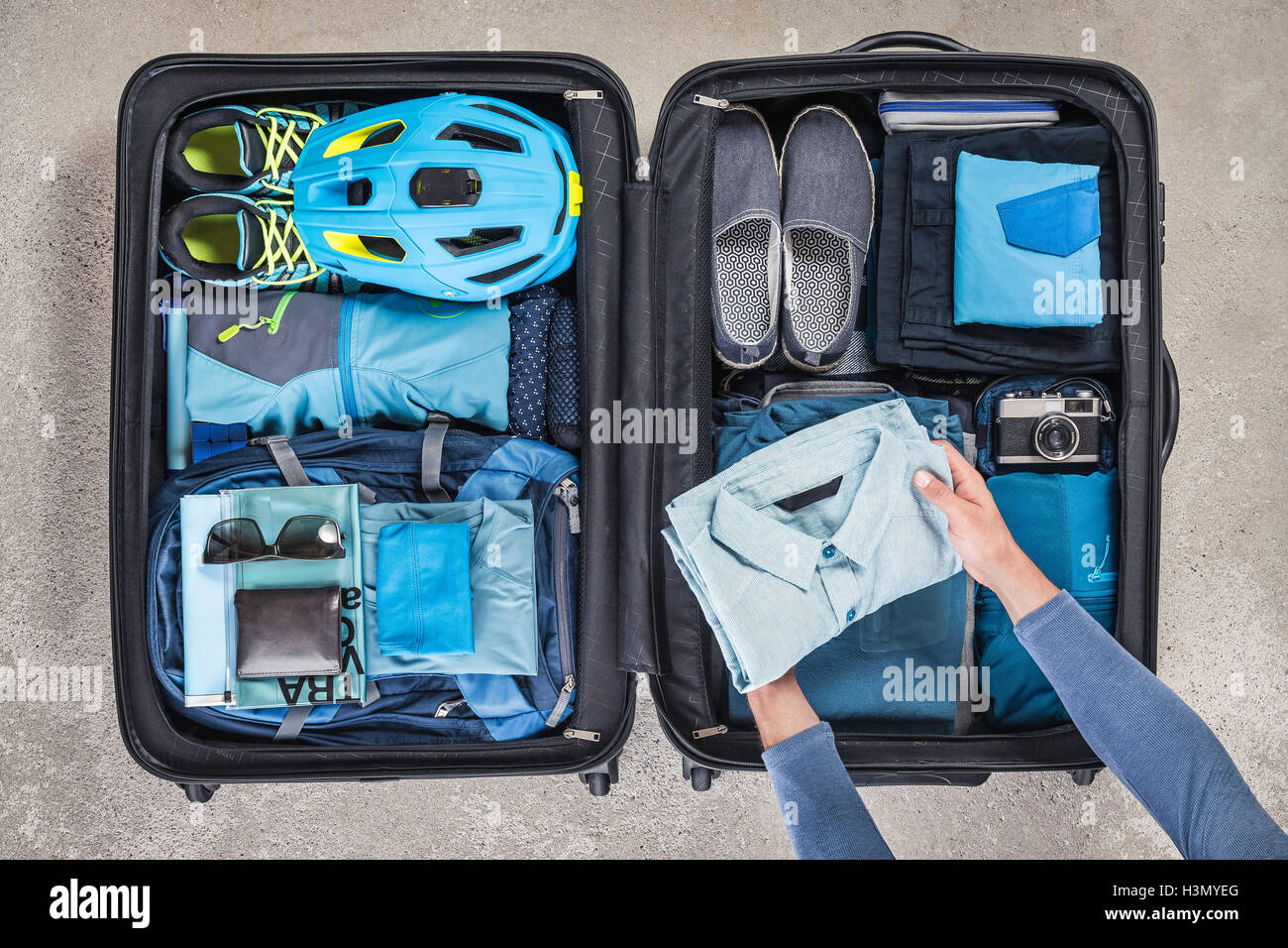 Vista aerea delle mani dell'uomo valigia di imballaggio con scarponi da trekking, bicicletta, casco, zaino, fotocamera retrò e maglietta blu Foto Stock