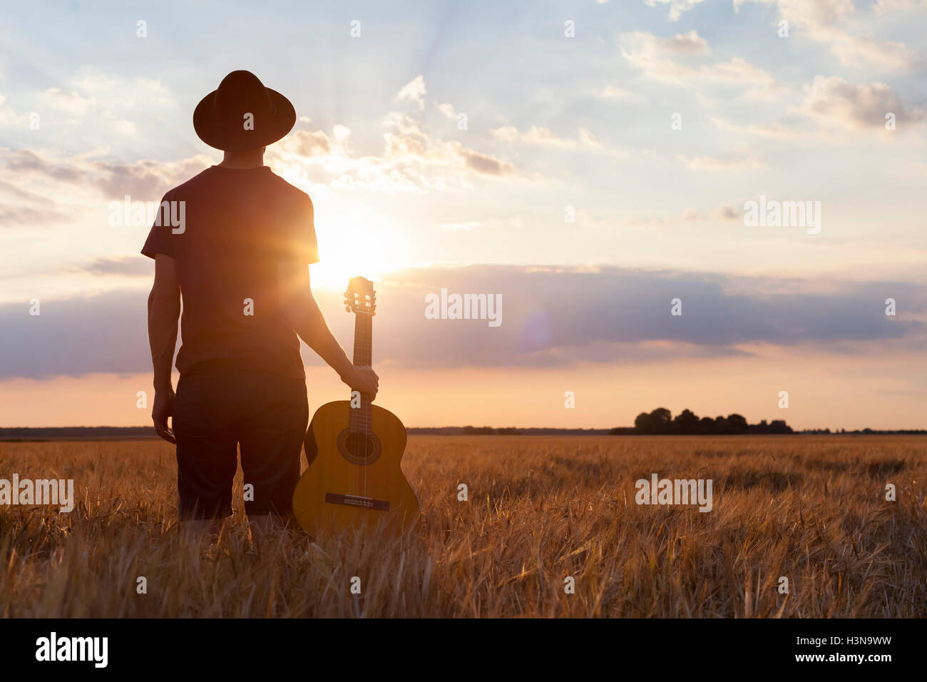 Musicista tenendo la chitarra acustica e camminare nei campi estivi al tramonto Foto Stock