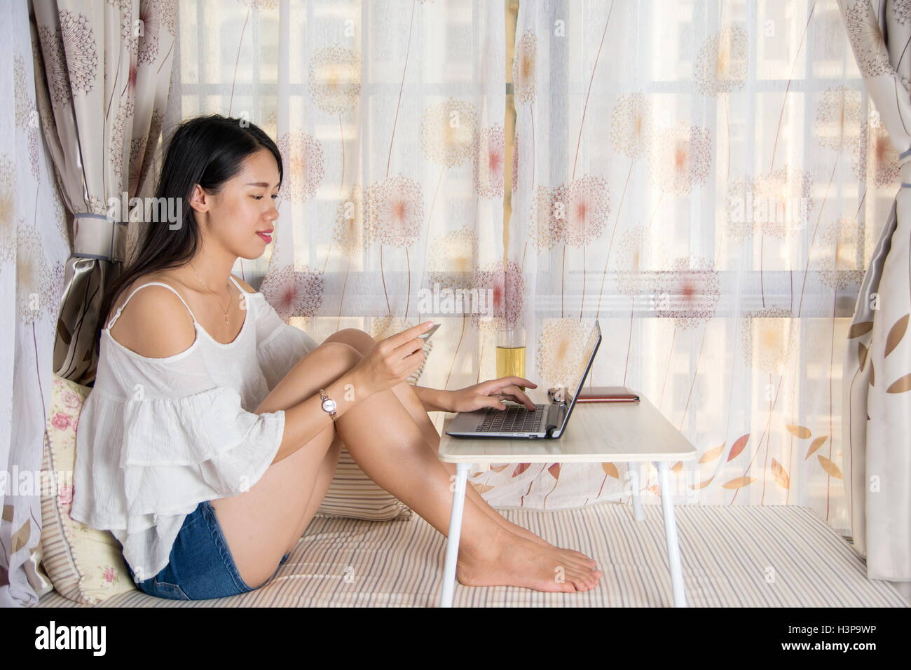 Giovane donna shopping online seduta mediante la finestra Foto Stock