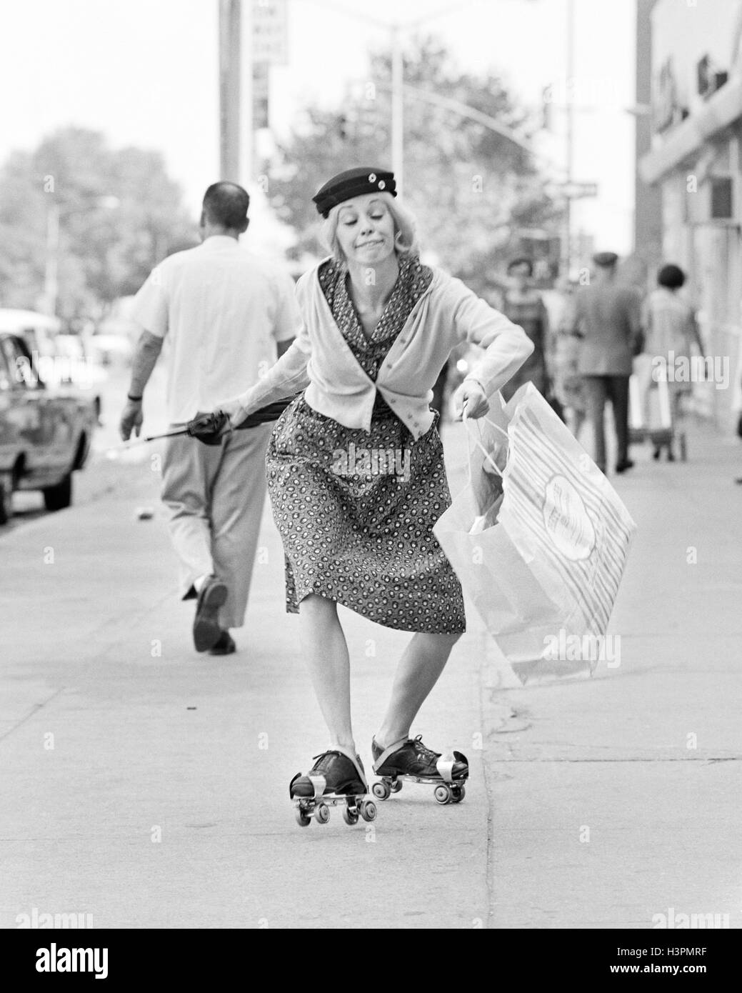 Anni sessanta anni settanta donna CARATTERE UNA SHOPPING BAG LADY divertente con espressione facciale indossando i pattini a rotelle pattinaggio sul marciapiede della città Foto Stock