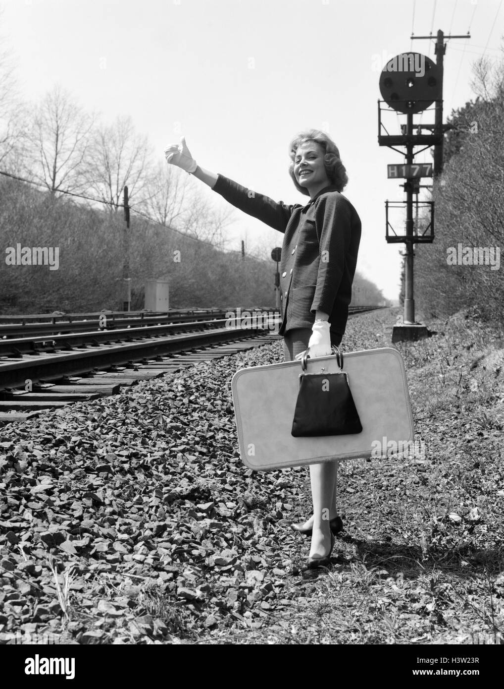 Anni Cinquanta anni sessanta donna sorridente tenendo la valigia in piedi accanto a binari mano guantata pollice in aria per l'attacco di una corsa Foto Stock