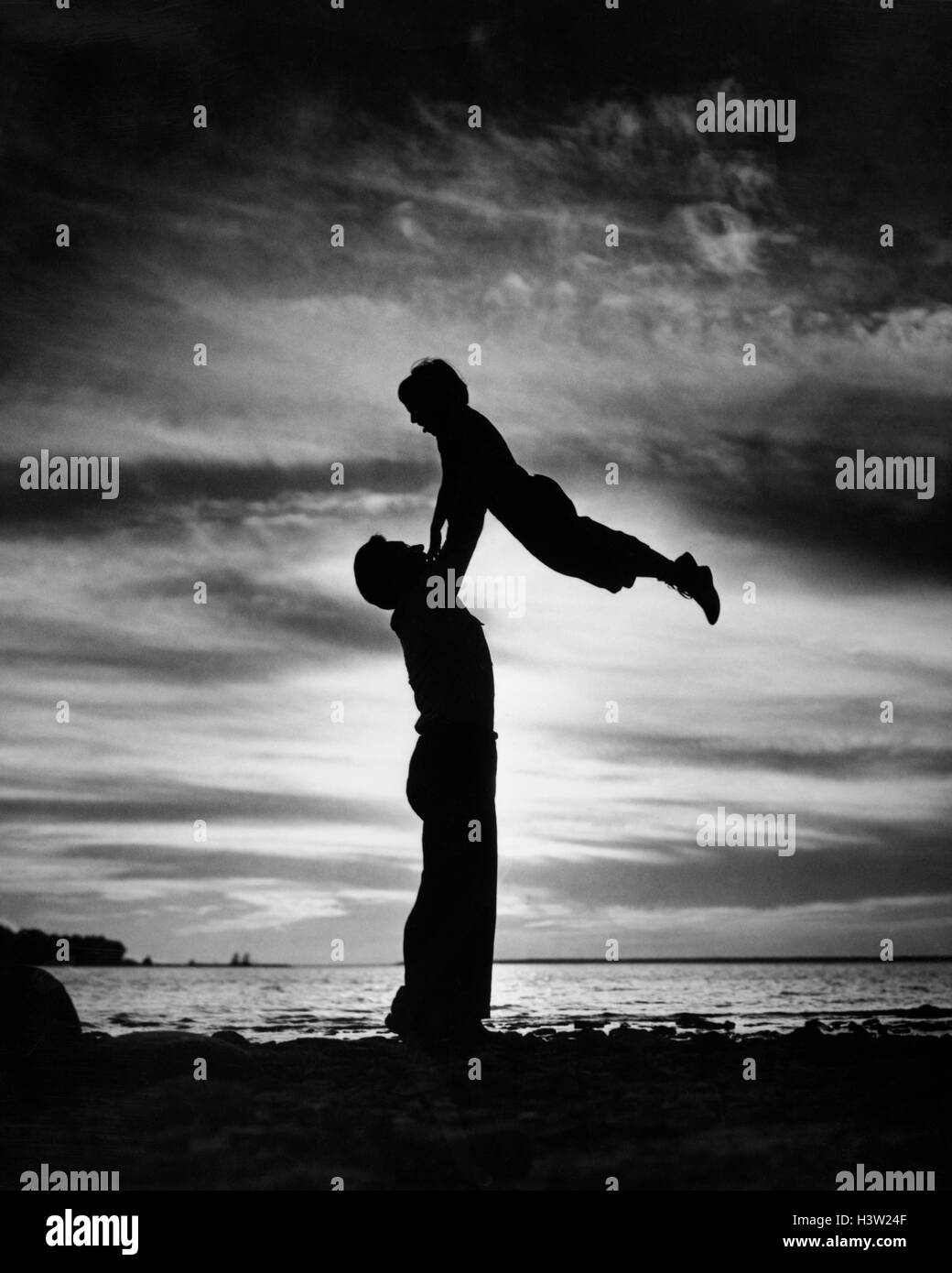Negli anni quaranta anni cinquanta anonimo stagliano uomo padre sollevamento figlio BAMBINO IN ARIA SULLA spiaggia al tramonto Foto Stock
