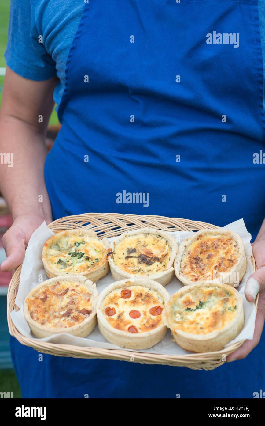 Un cestello di sei quiche con vari ripieni salati detenute da uomo in grembiule blu ritagliato dal collo in giù con spazio di copia Foto Stock