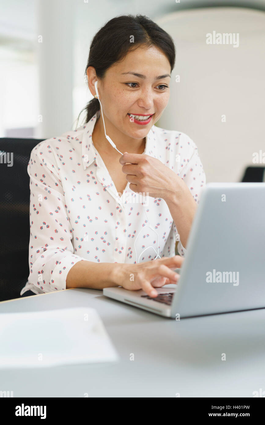 Sorridente imprenditrice utilizzando le cuffie mentre si lavora sul computer portatile alla scrivania in ufficio Foto Stock