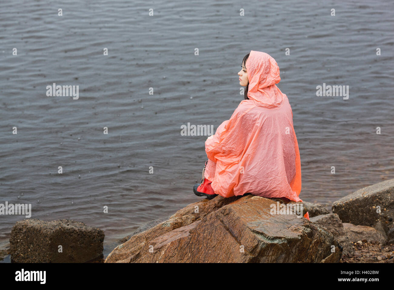 Premurosa donna che indossa un impermeabile seduta su roccia sul lungolago durante la stagione delle piogge Foto Stock