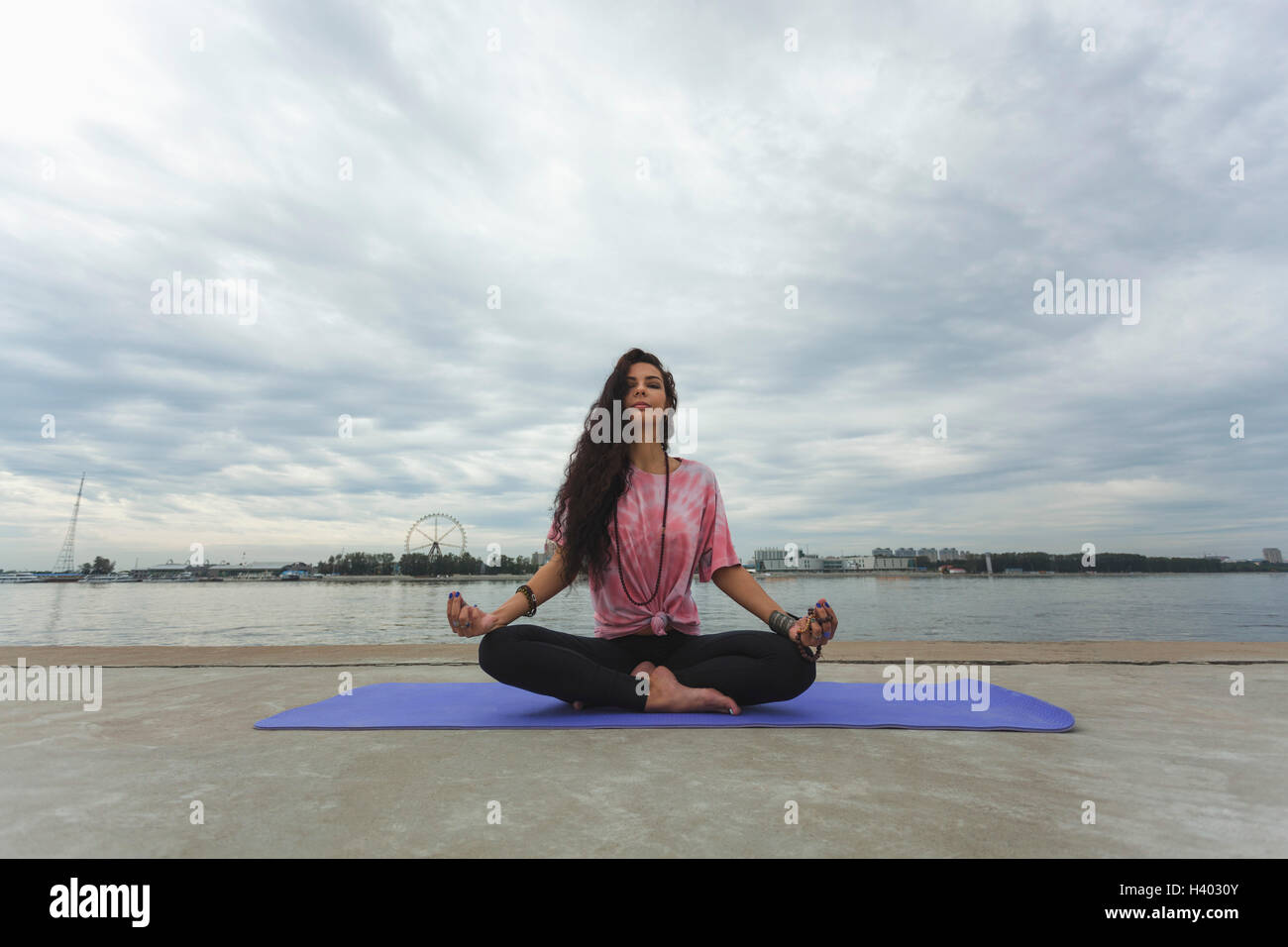 Donna meditando nella posizione del loto a riverbank contro il cielo nuvoloso Foto Stock