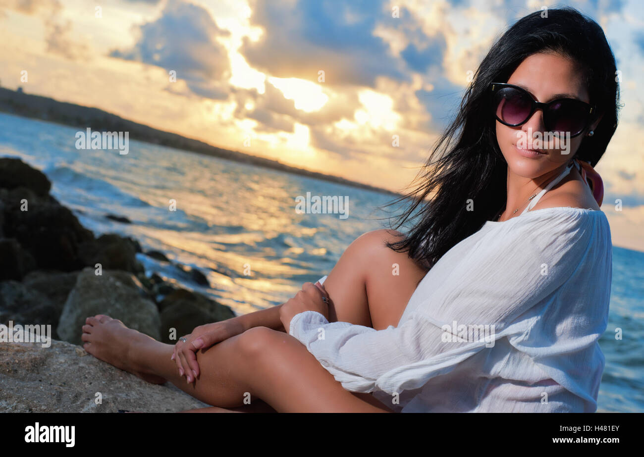 Giovane e bella bruna donna con occhiali da sole seduto su di una roccia  dall'oceano al tramonto Foto stock - Alamy