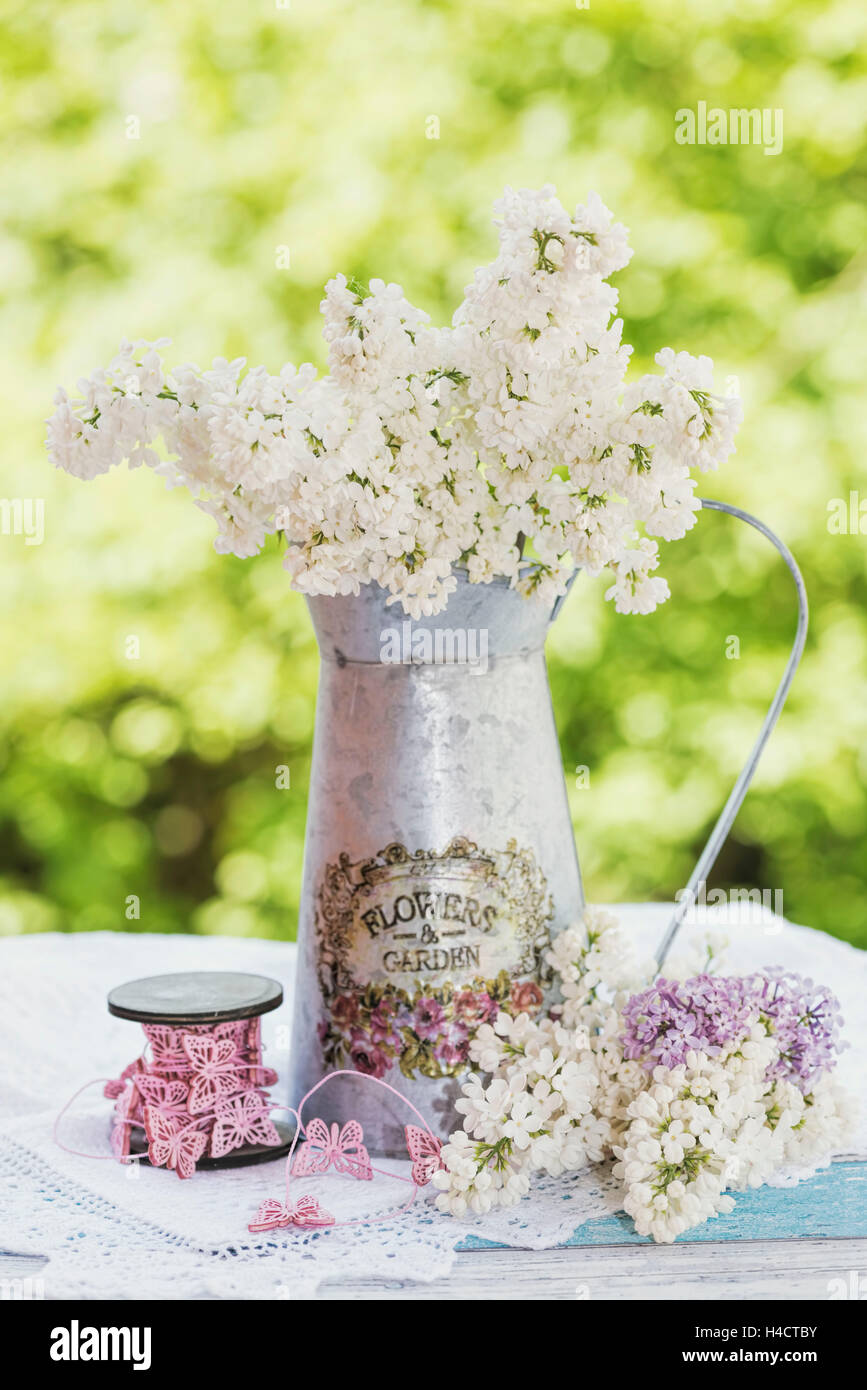 Brocca in metallo bianco con il lilla, bianco cappelli superiori su una vecchia tavola di legno, ruolo con farfalla il cavo singolo, fiori per decorazione, Foto Stock