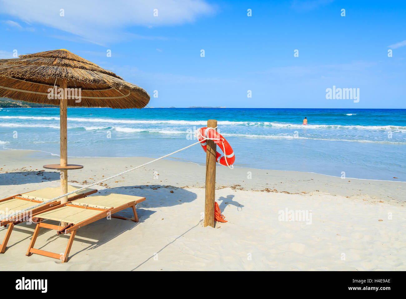 Sdraie con ombrelloni e rosso anello di vita appesi ad un palo di legno con un misterioso giovane uomo a nuotare in un mare azzurro acqua in background, l'isola di Sardegna, Italia Foto Stock