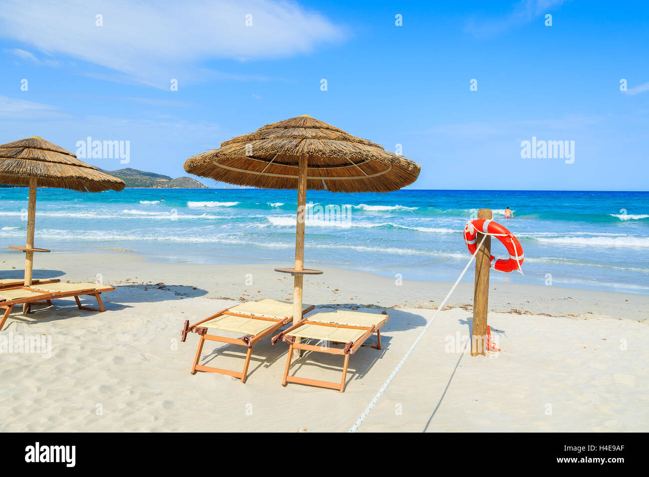 Sdraie con ombrelloni e rosso anello di vita appesi ad un palo di legno con un misterioso giovane uomo a nuotare in un mare azzurro acqua in background, l'isola di Sardegna, Italia Foto Stock