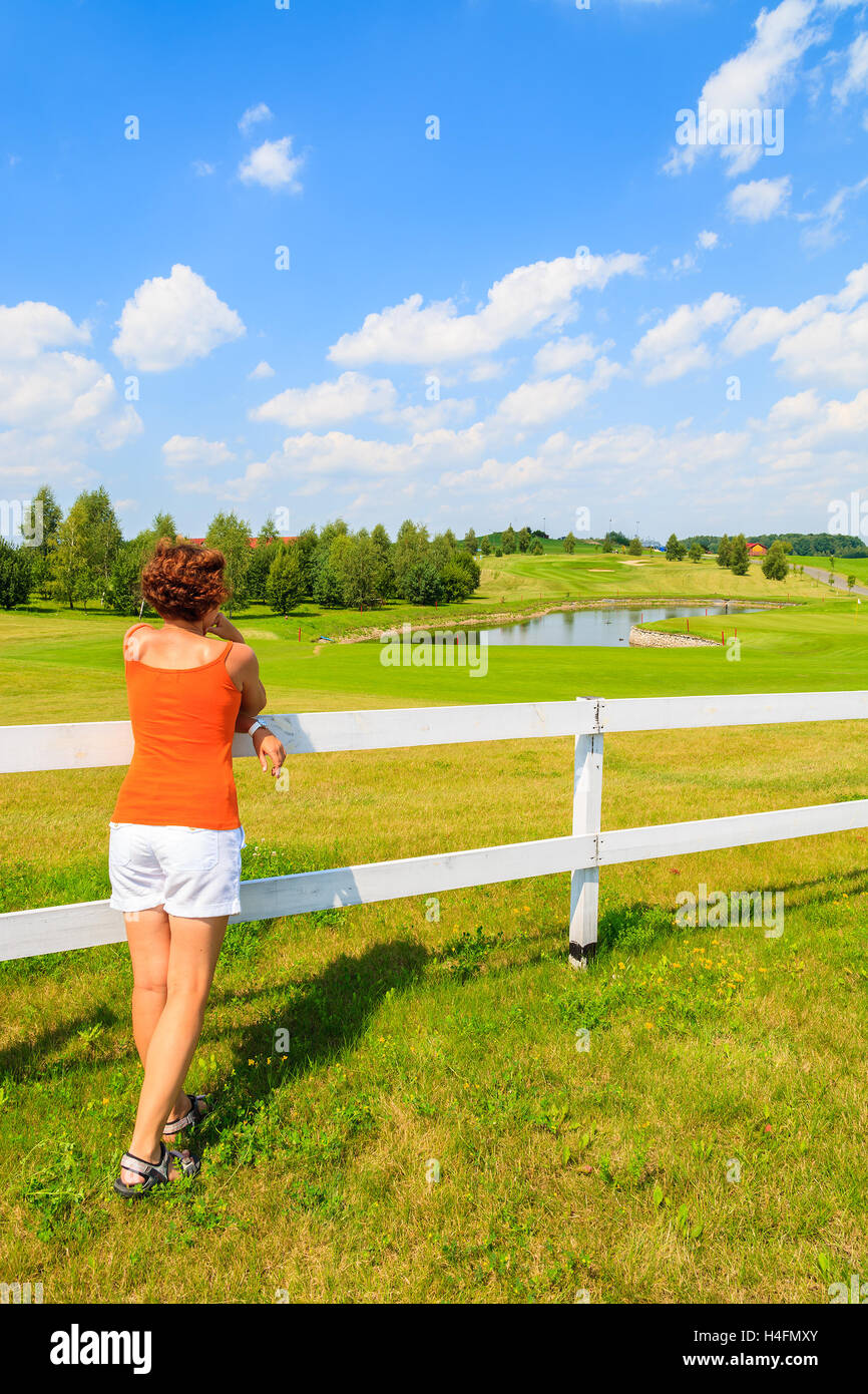 Giovane donna si erge contro un recinto bianco e guarda al golf club area su soleggiate giornate estive, Polonia Foto Stock